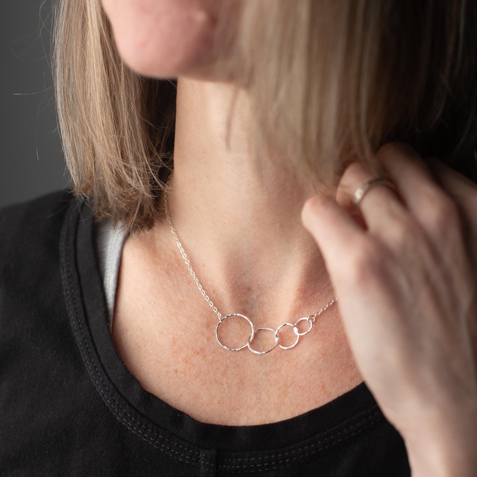 A close-up lifestyle shot of a woman wearing a delicate silver necklace with four interlocking textured circular rings. The necklace is displayed on her neck, with her hand resting near her collarbone, wearing a black top against a dark gray background.