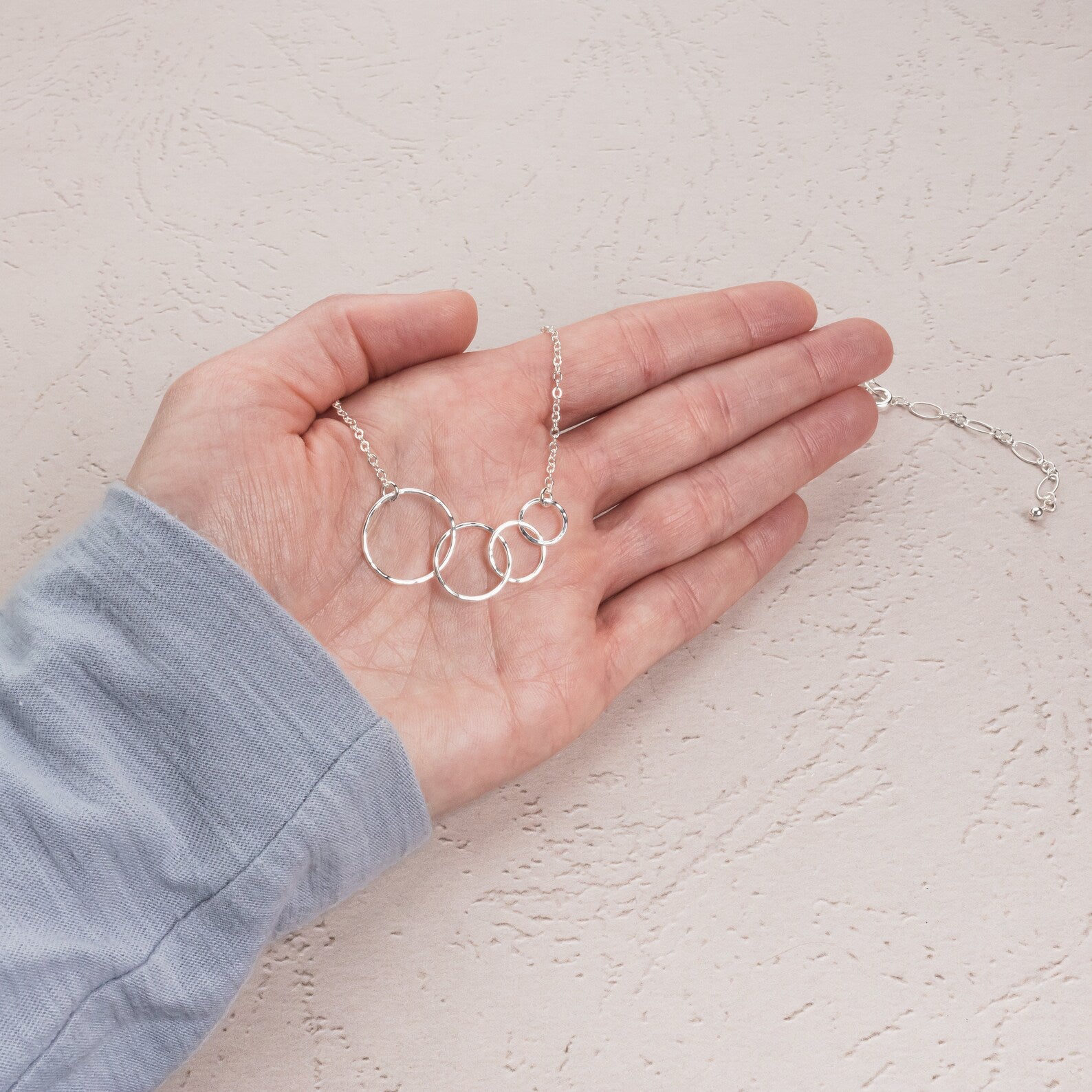 A silver necklace with four interlocking open rings on a delicate chain, held in a person's hand against a textured off-white background.