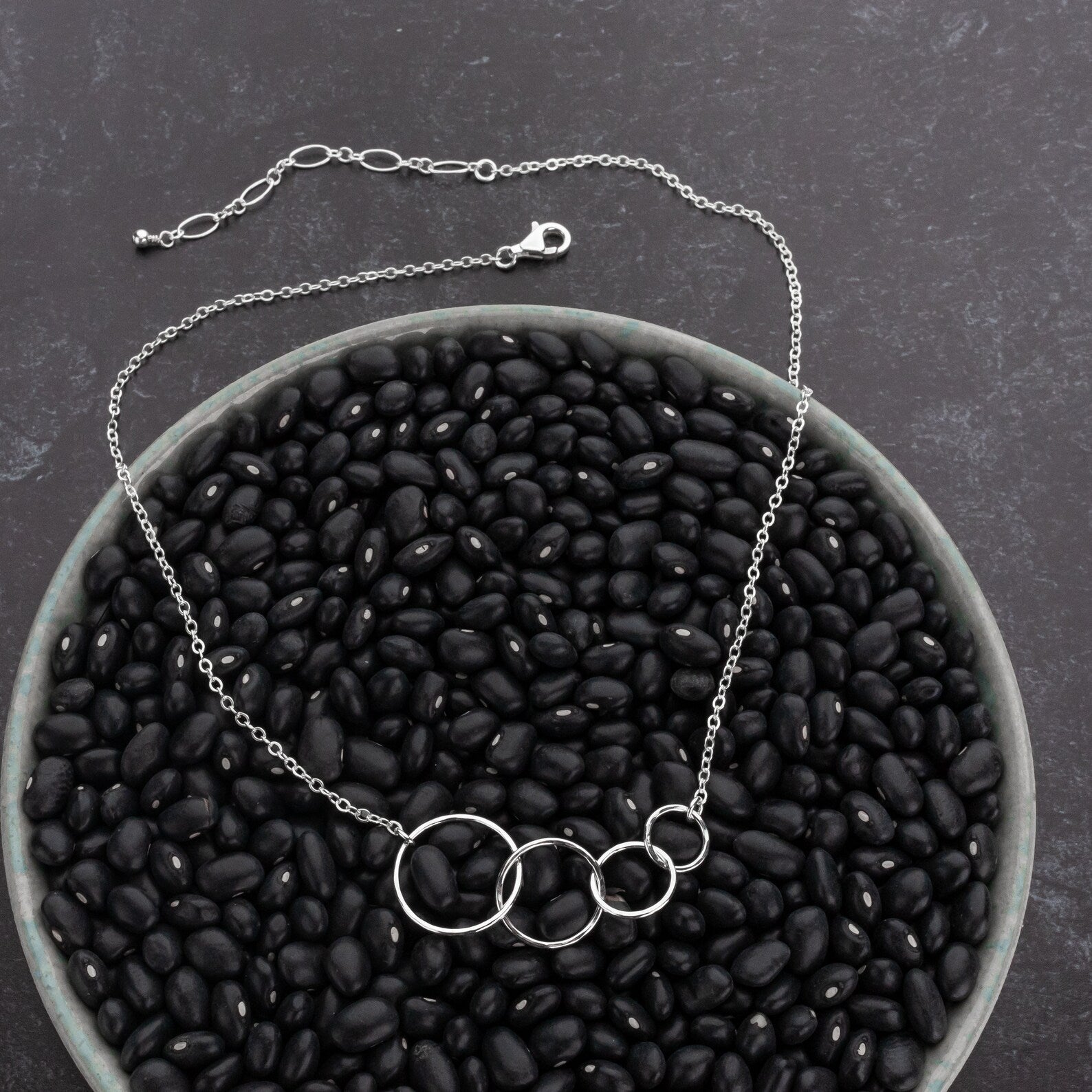 A silver chain necklace with four interlocking circular rings, displayed on a bed of black beans inside a gray bowl against a dark textured background.