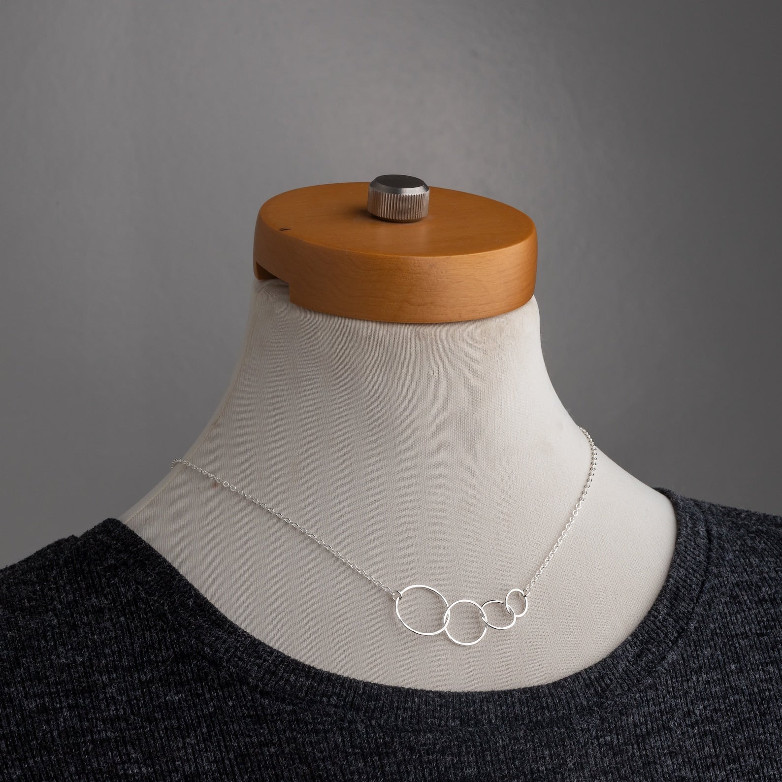 A delicate silver necklace with four interlocking open circles displayed on a cream-colored mannequin bust wearing a dark gray knit top, set against a neutral gray background.