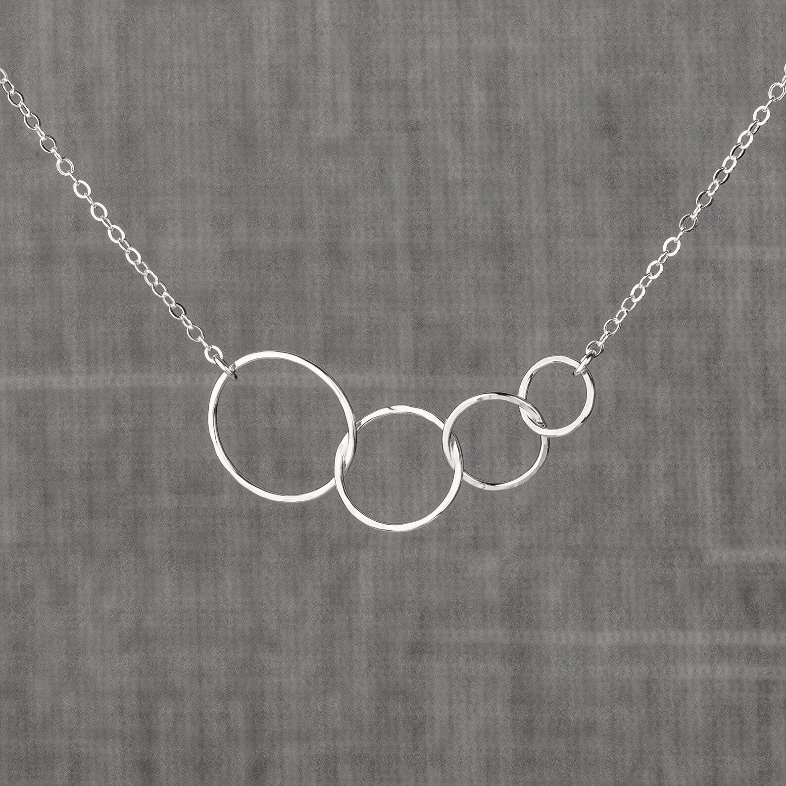 A silver necklace with four interlocking open circles of decreasing size on a delicate chain, displayed against a textured gray fabric background.