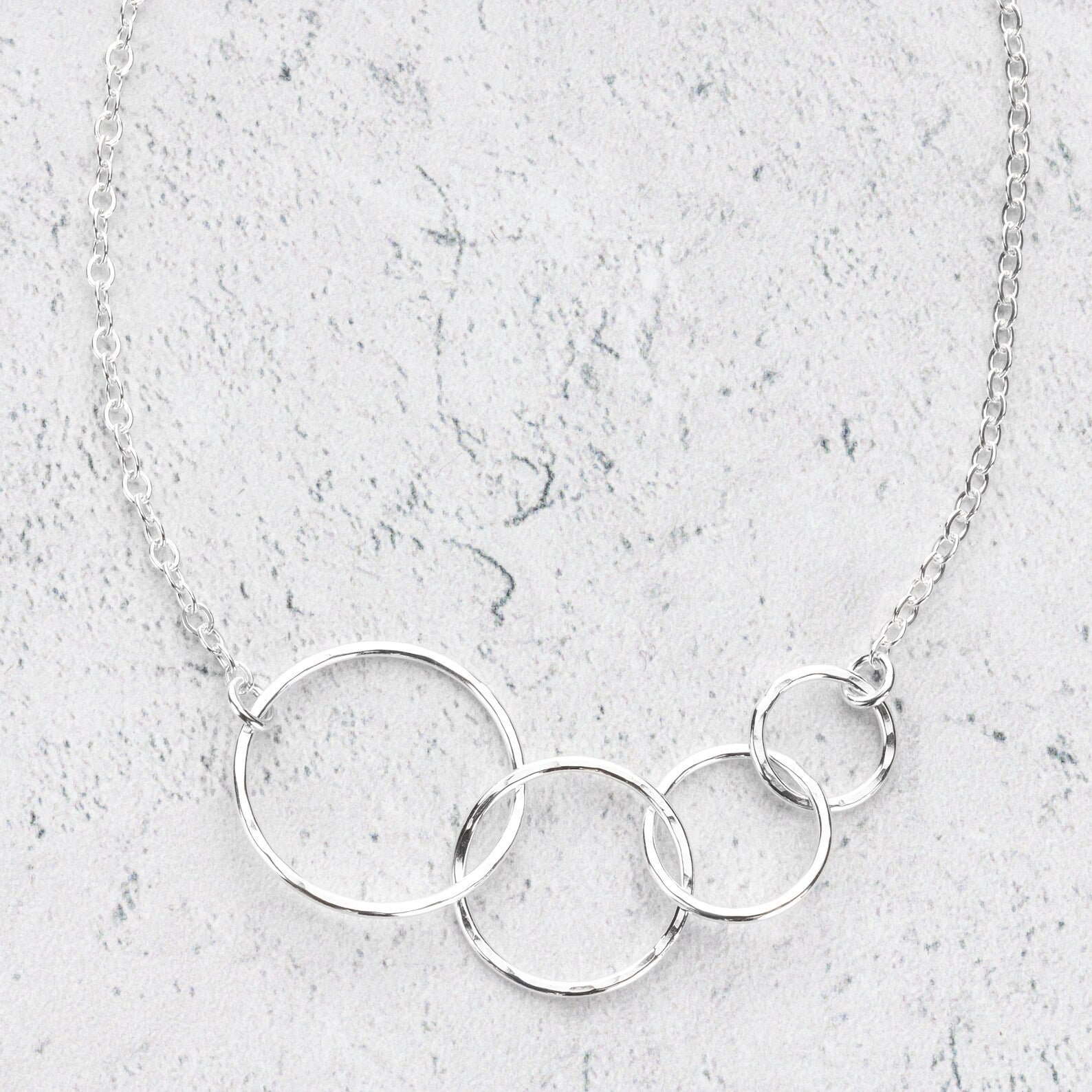A silver necklace featuring interlocking circles of varying sizes on a delicate link chain, displayed on a textured light gray stone surface.