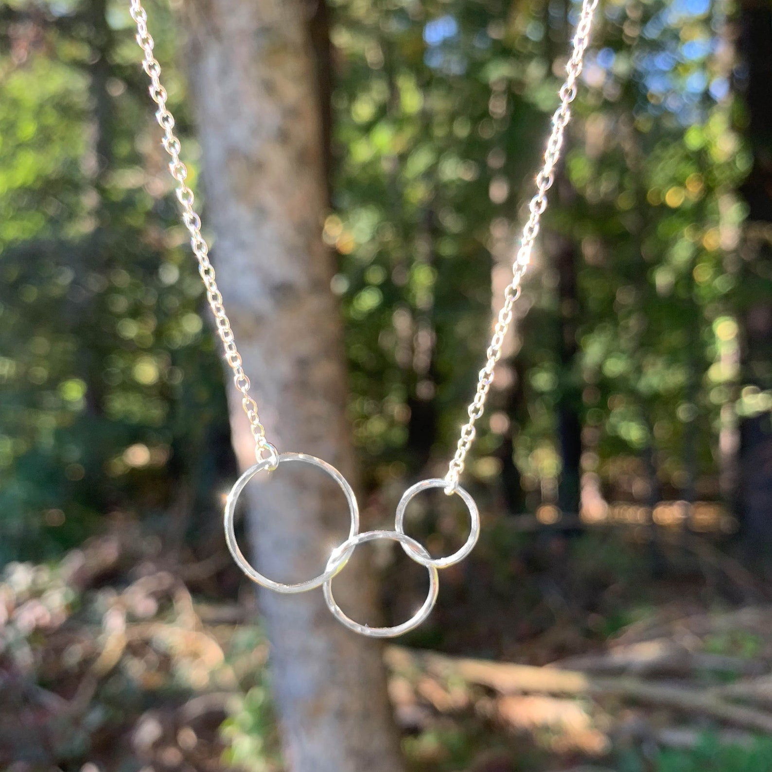 A silver necklace with three interlocking circular rings on a delicate chain, photographed outdoors against a blurred forest background.