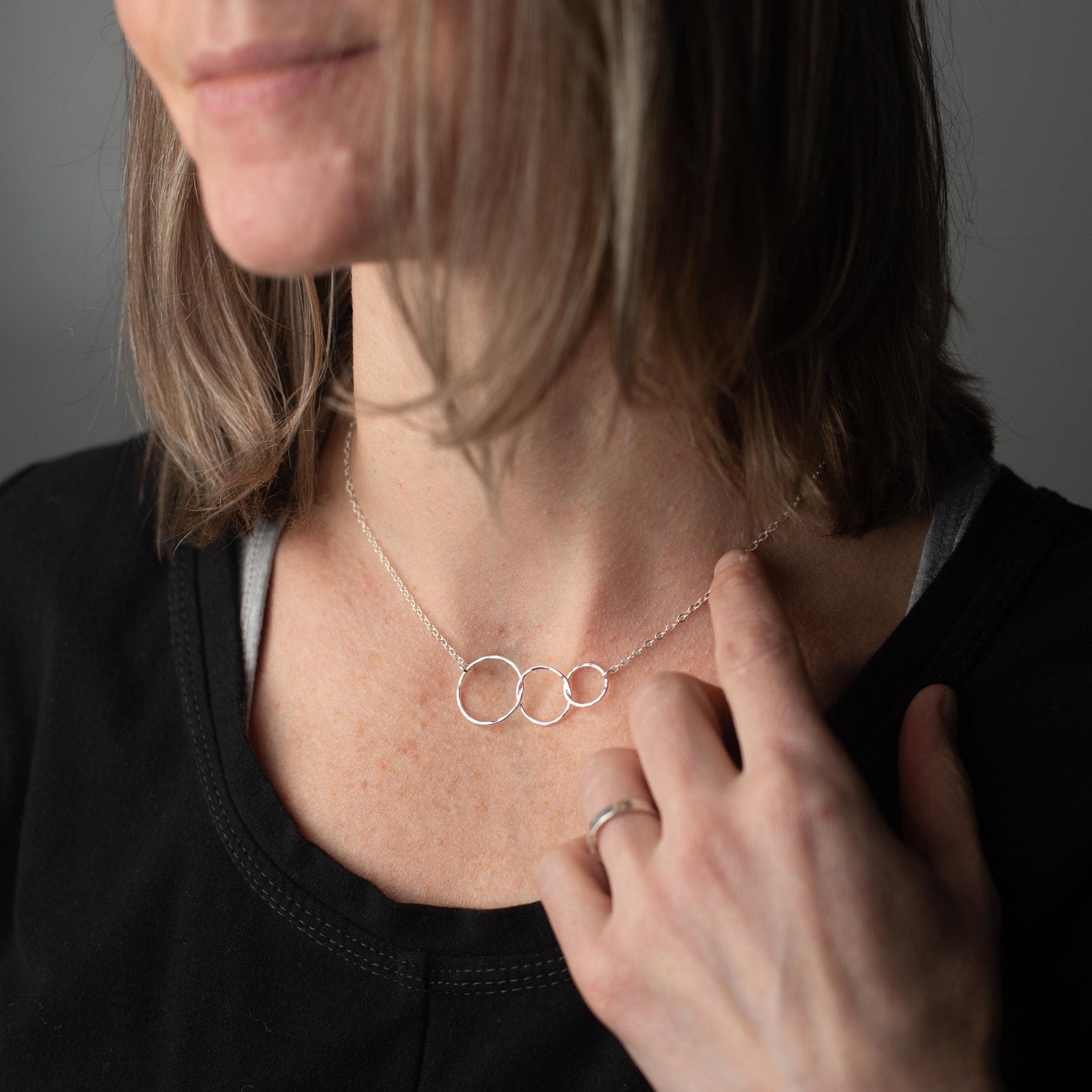 A close-up of a woman wearing a delicate silver necklace with three interlocking open rings, touching the pendant with her finger. She is wearing a black top against a neutral gray background.