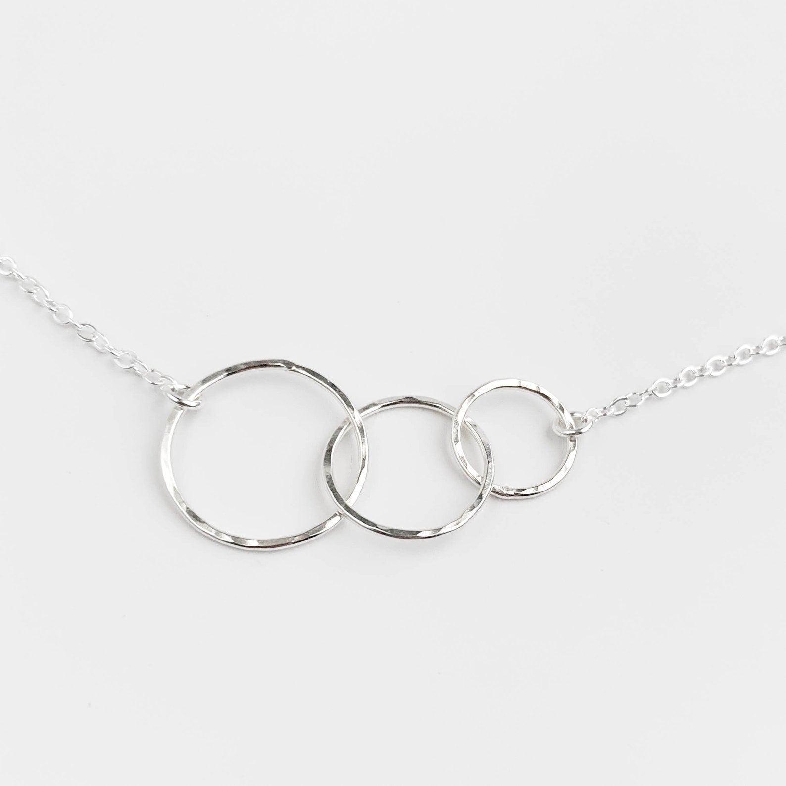 A silver necklace featuring three interlocking textured circular rings on a delicate chain, photographed against a clean, light gray background.