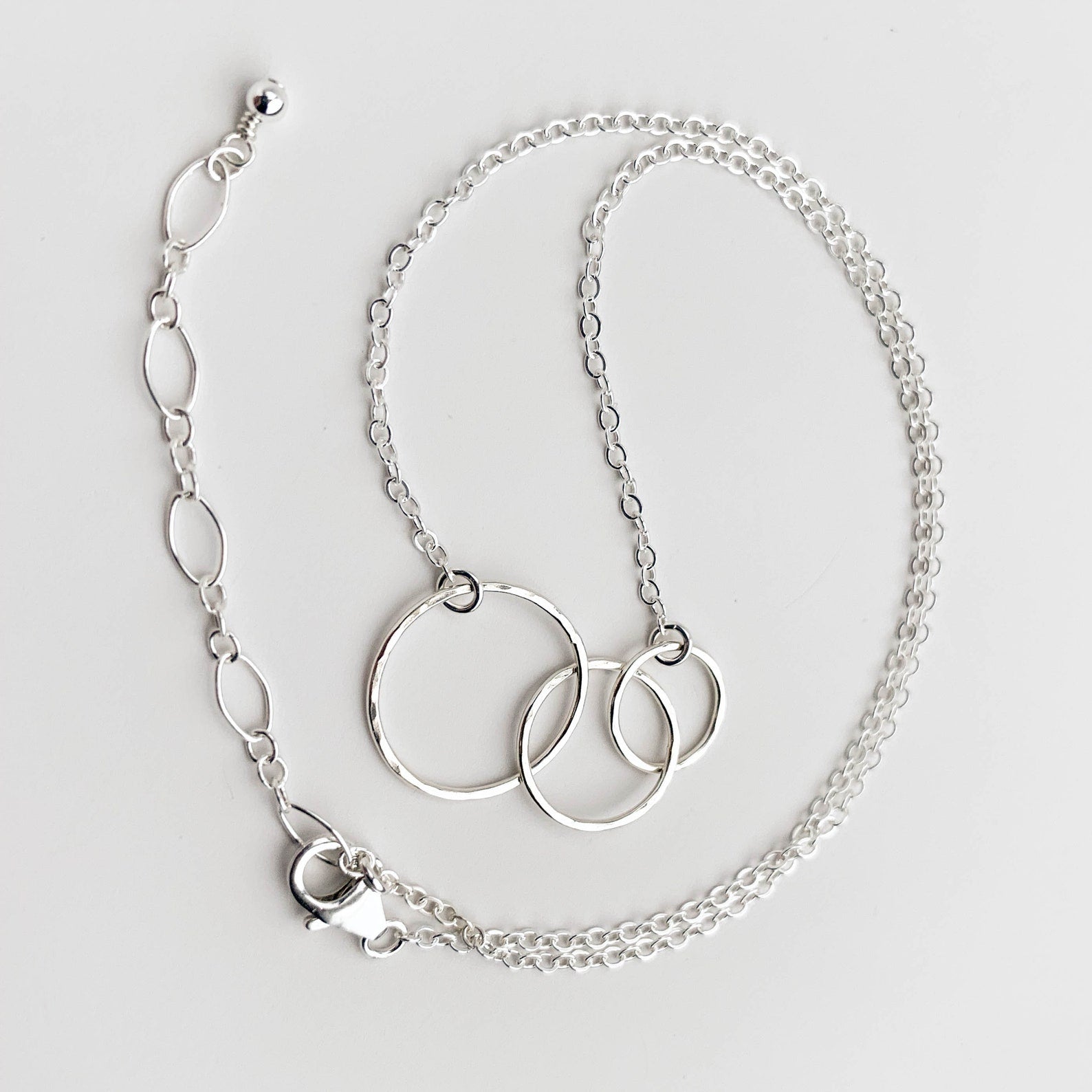 A silver chain necklace featuring three interlocking textured circular rings, displayed on a clean white background.