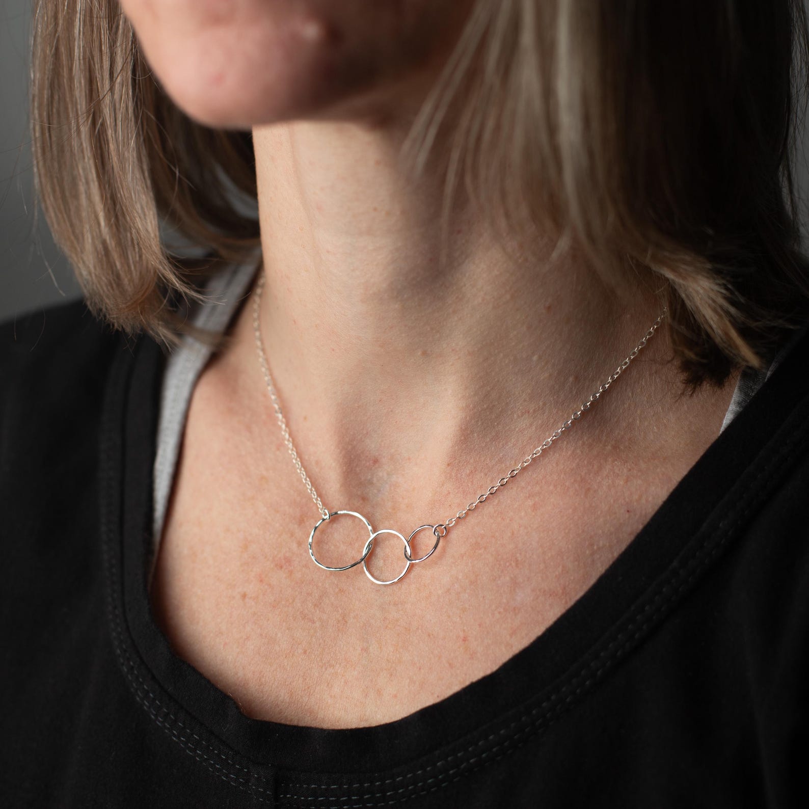 A close-up of a woman wearing a delicate silver necklace with three interlocking open rings at the center, styled with a black top.