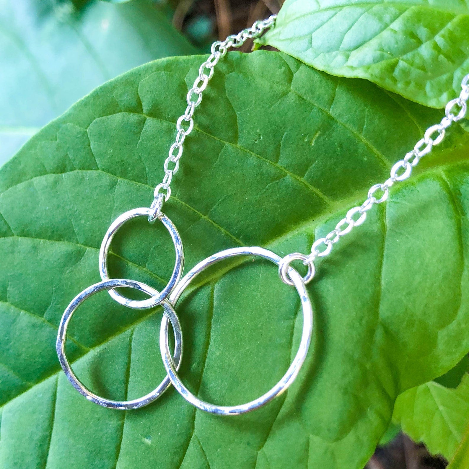 A silver necklace with three interlocking open circular rings on a delicate chain, displayed on a large green leaf.