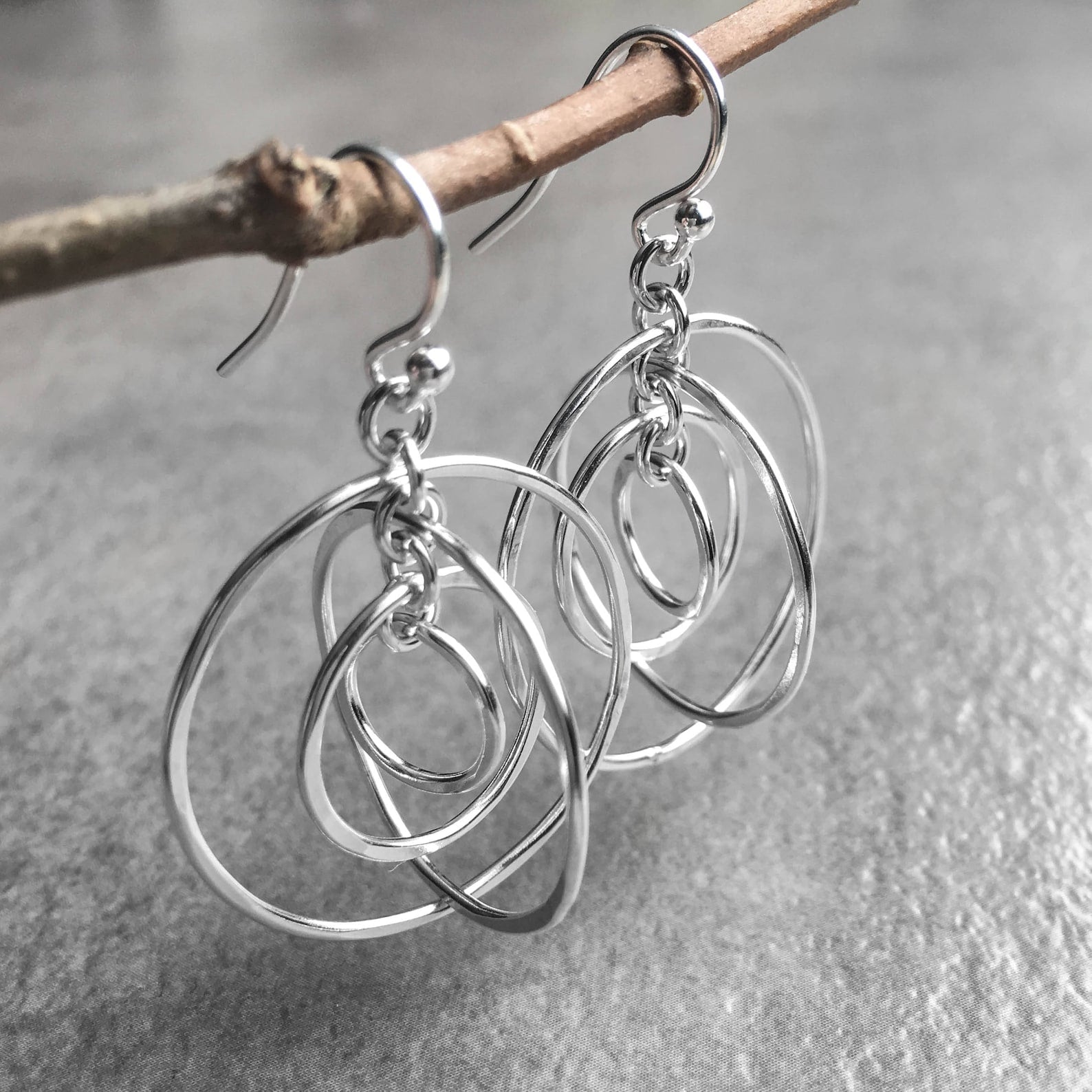 Sterling silver dangle earrings with interlocking multi-ring circles, hanging from a small natural twig against a textured gray background.
