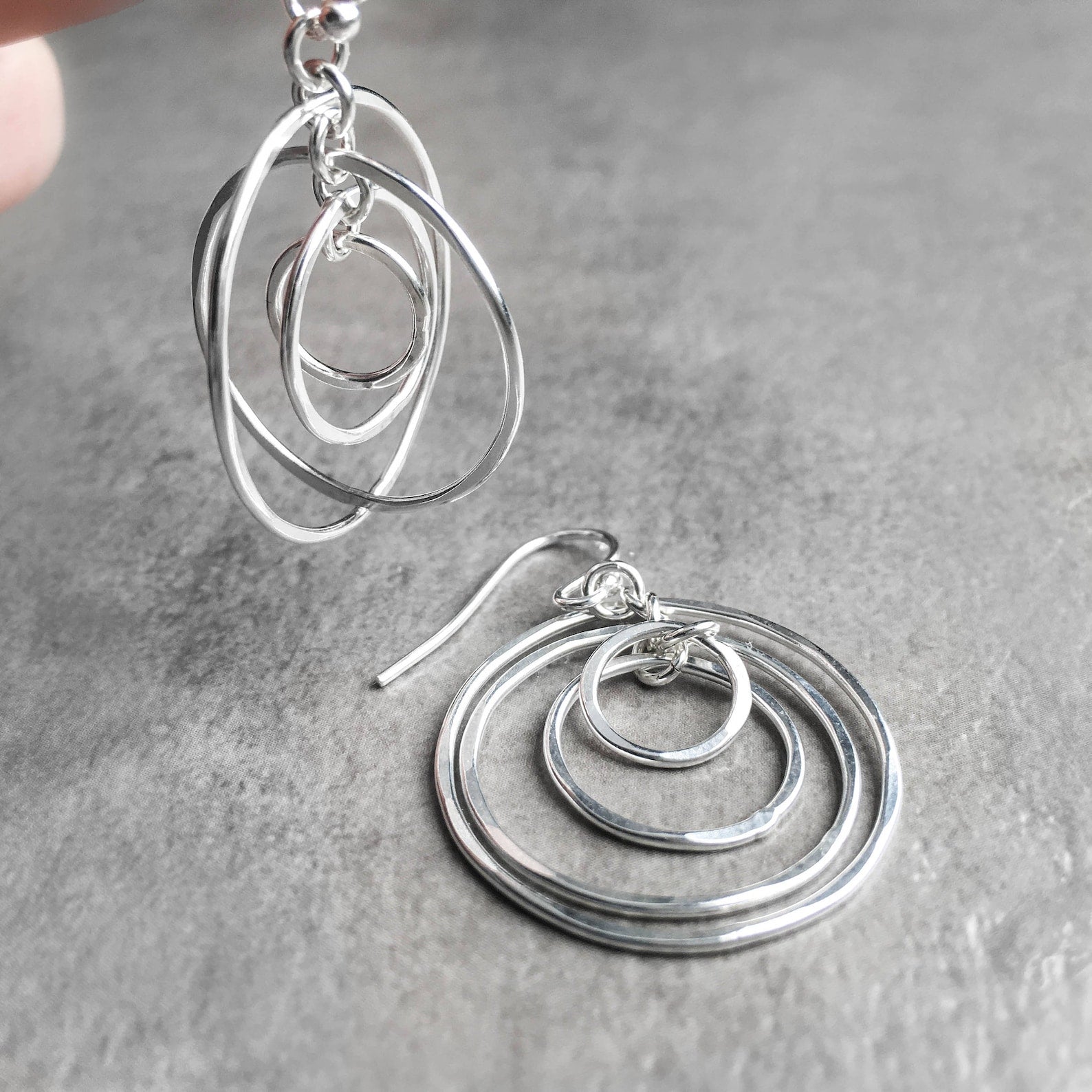 Sterling silver dangle earrings with nested concentric circle designs, shown on a textured gray surface with a hand partially visible holding one earring.