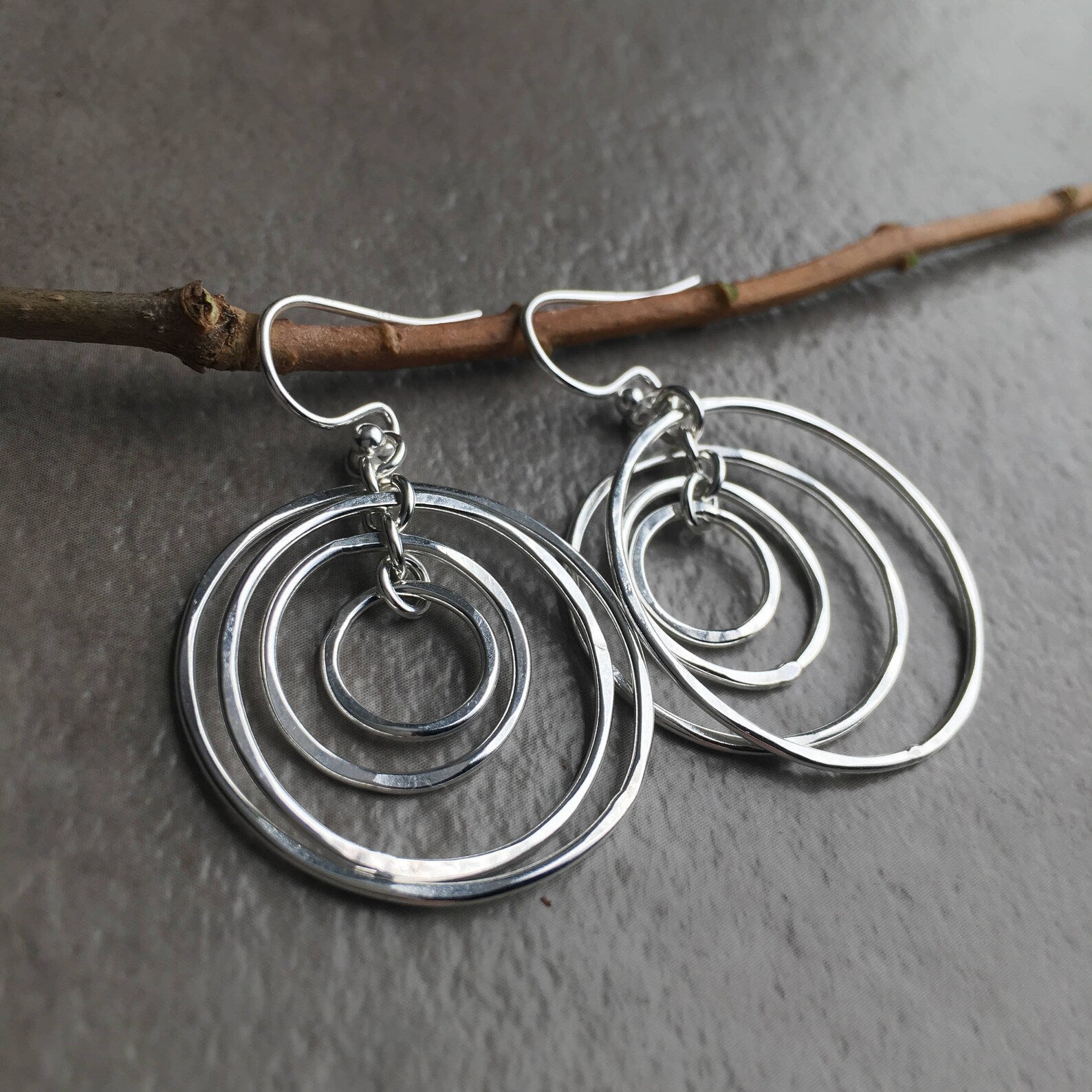 A pair of silver dangle earrings with nested, openwork concentric circles, hanging from fishhook wires and displayed on a textured gray surface with a small brown twig behind them.