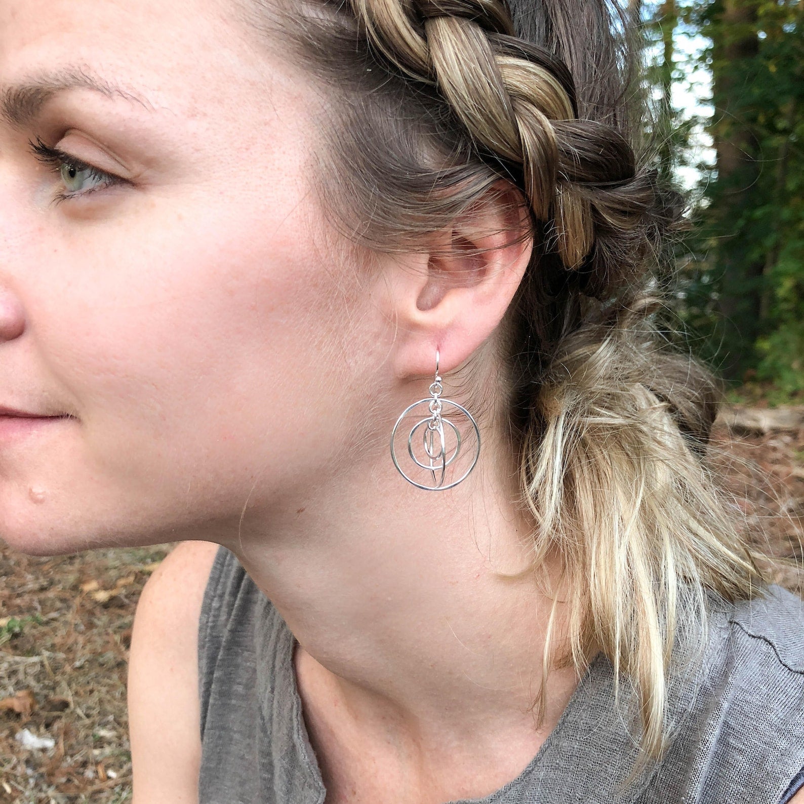 A close-up side profile of a woman wearing a silver dangle earring with interlocking circular wire details, set against an outdoor background of trees and ground cover.