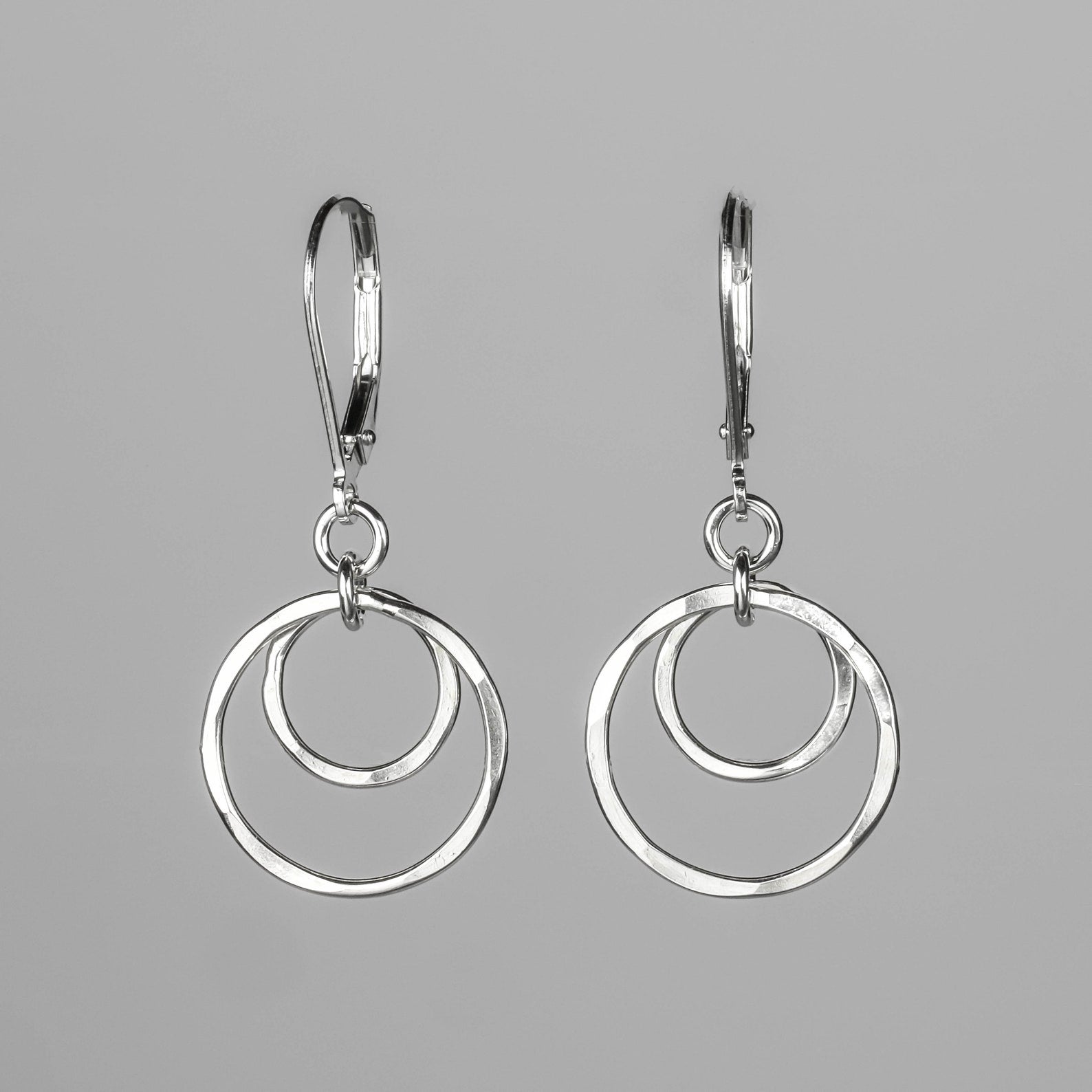 A pair of silver drop earrings with a double-ring concentric circle design, suspended from leverback hooks against a solid gray background.
