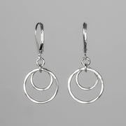 A pair of silver drop earrings with a double-ring concentric circle design, suspended from leverback hooks against a solid gray background.