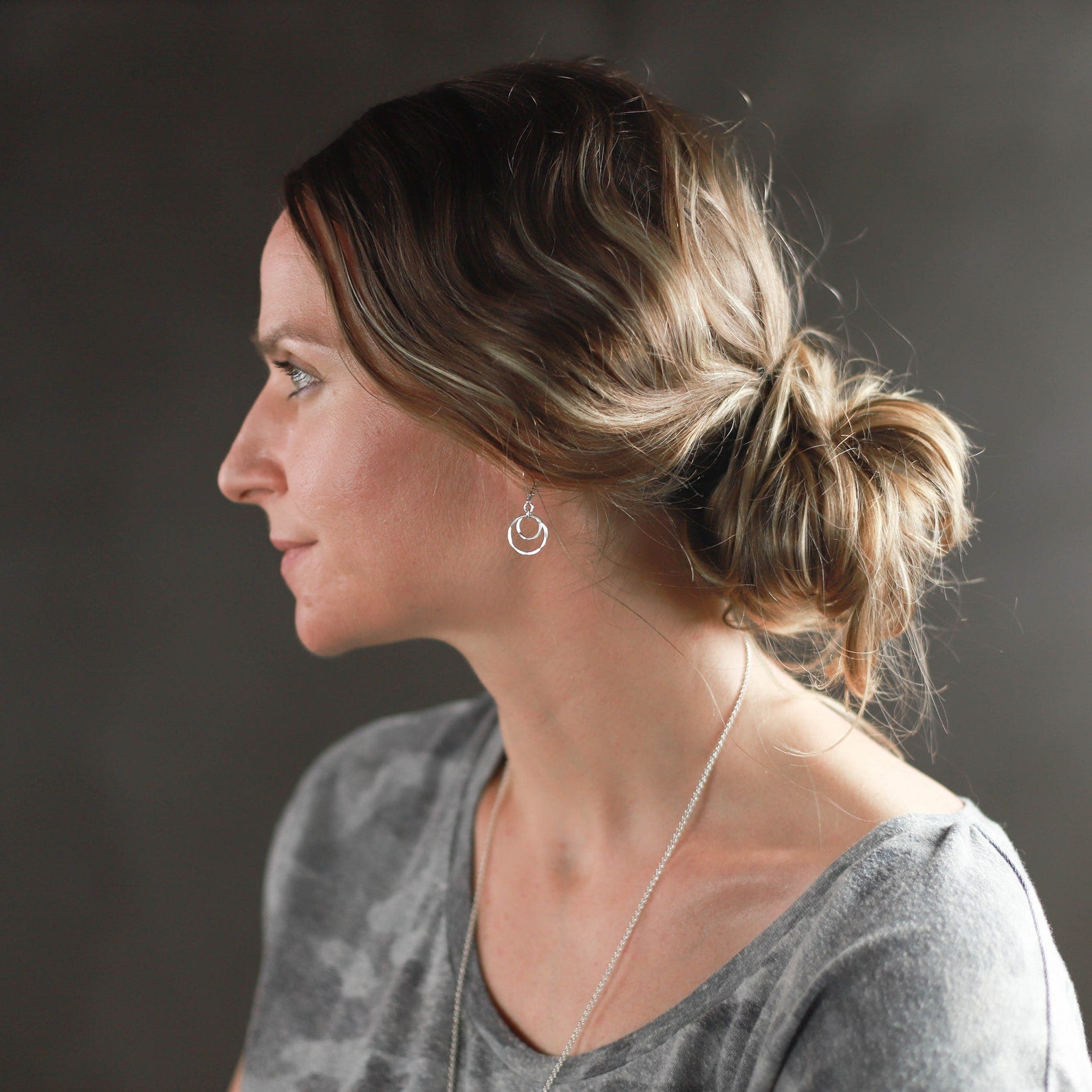 A woman in profile wearing a silver double-ring earring and a delicate silver chain necklace, styled in a messy bun, against a dark gray background.