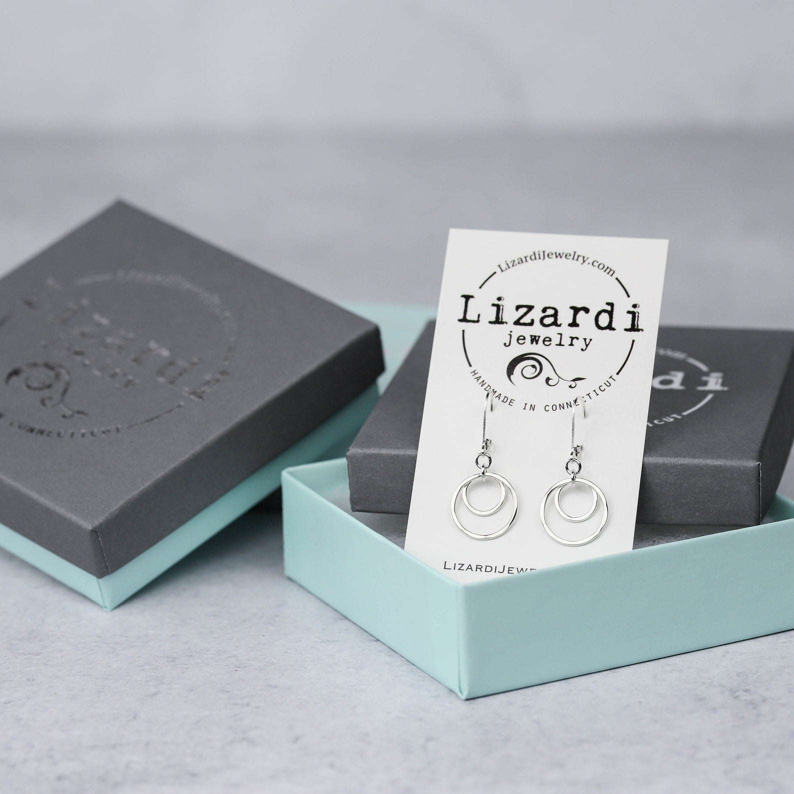 Sterling Silver double-circle earrings displayed on a white Lizardi Jewelry brand card, set inside an open light blue jewelry box on a light gray surface.