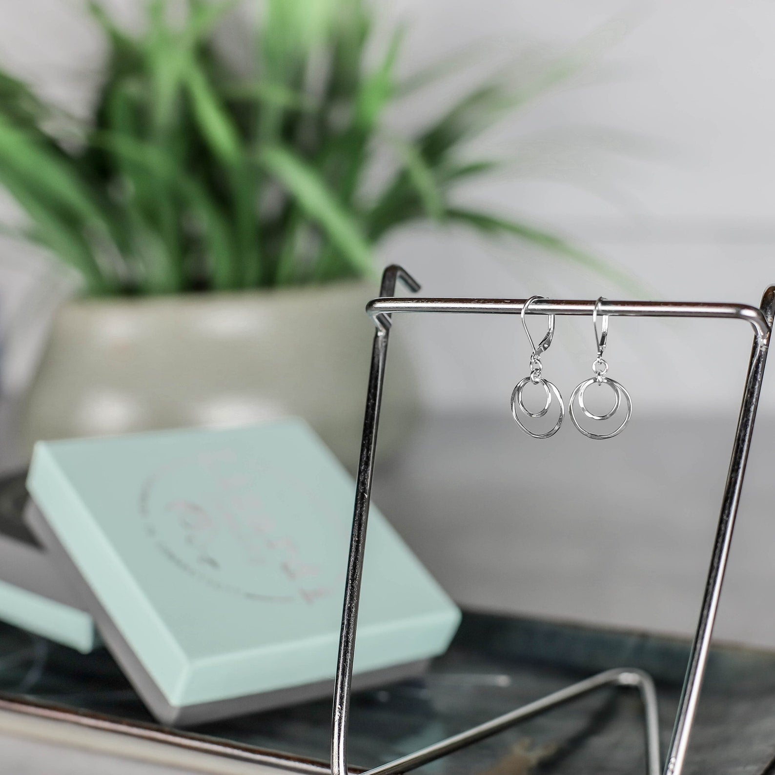 A pair of silver hoop drop earrings hanging from a metal display stand, with a mint jewelry box and a blurred green plant in the background.