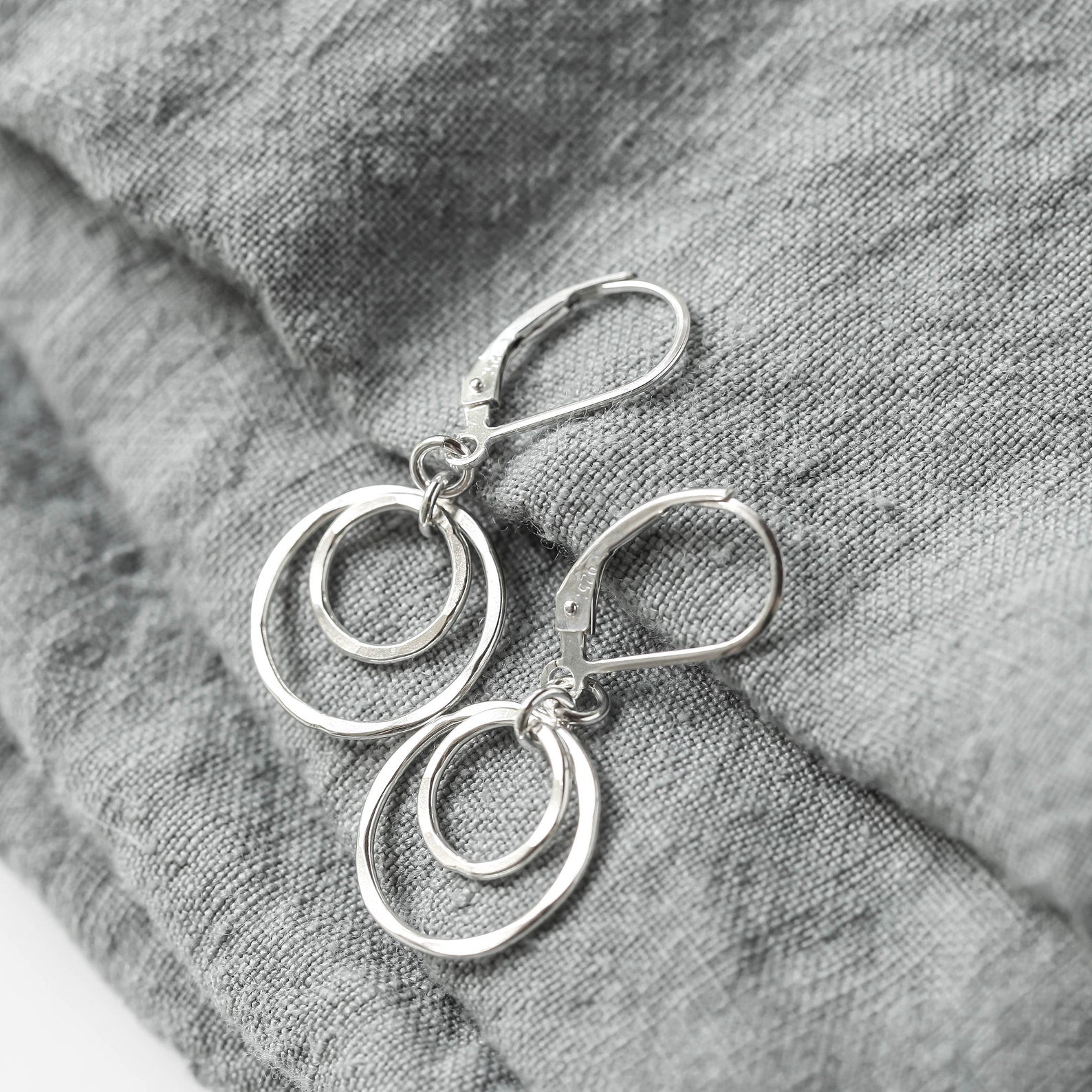 A pair of silver double-ring dangle earrings with leverback closures, displayed on a textured gray linen fabric background.