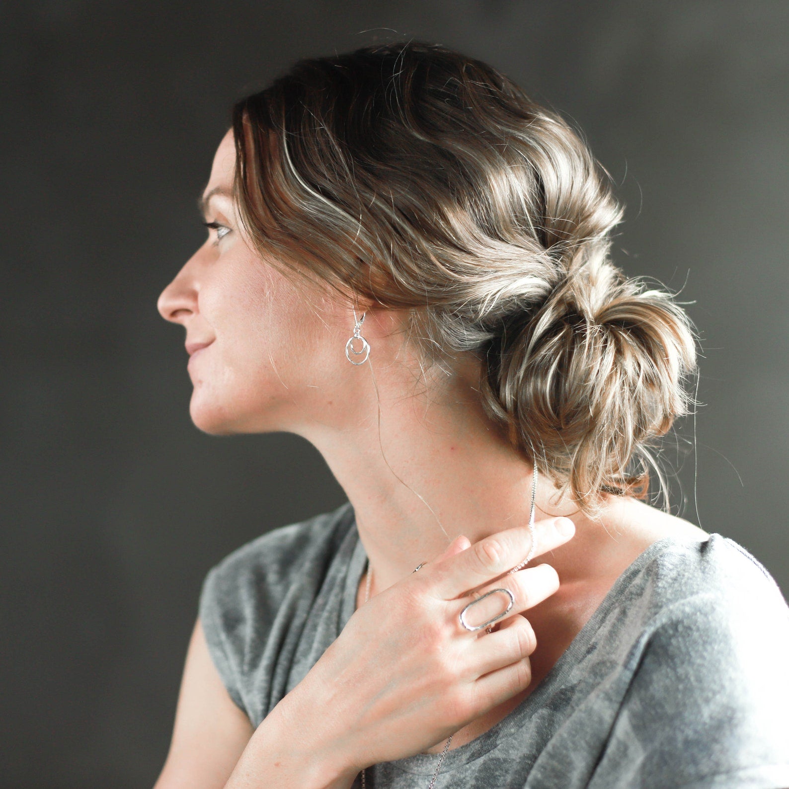 A woman in profile wearing delicate silver hoop earrings and a matching silver ring, with her hair styled in a low bun, against a dark gray background.