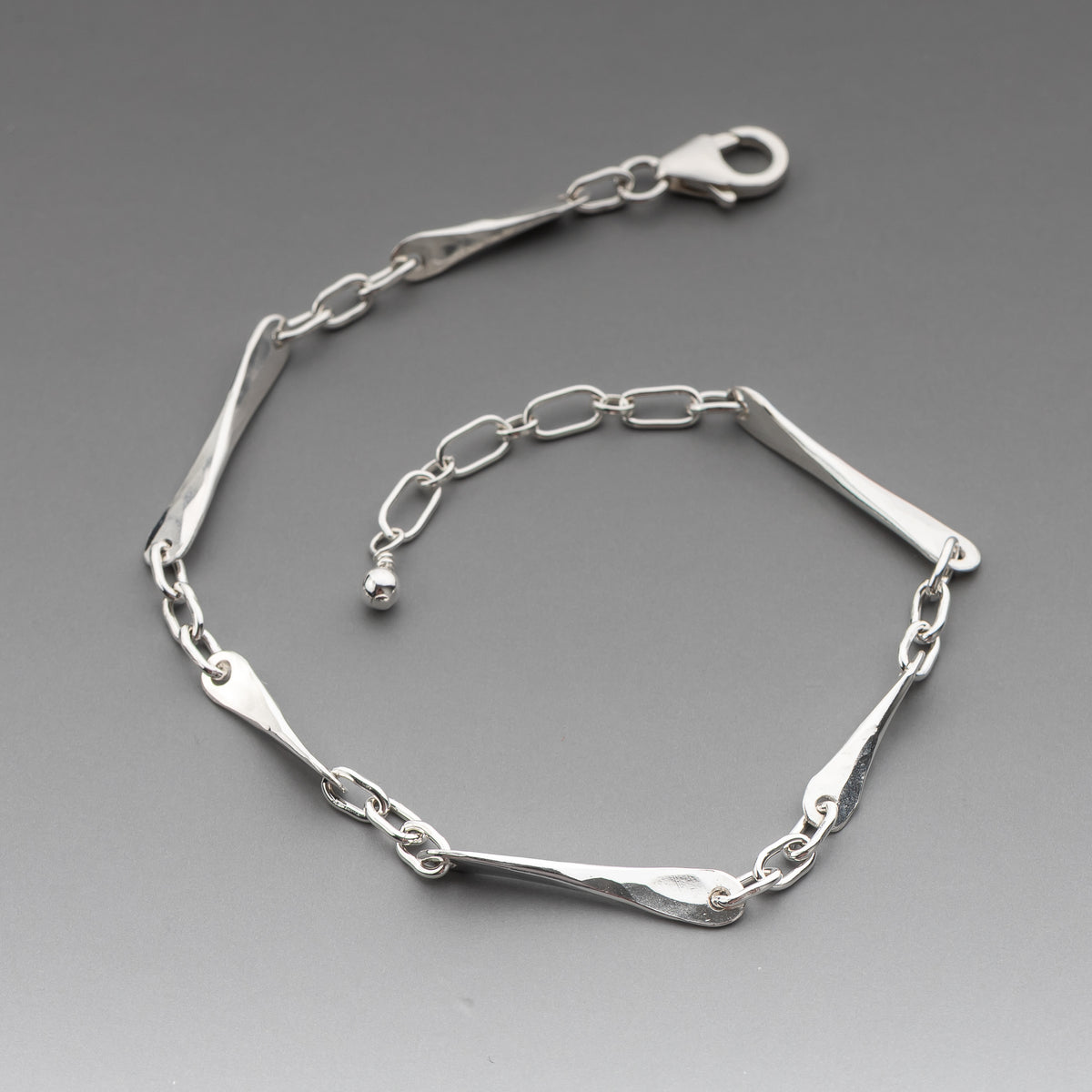 A polished sterling silver link bracelet with elongated hand hammered links and a lobster clasp, displayed on a neutral gray background.