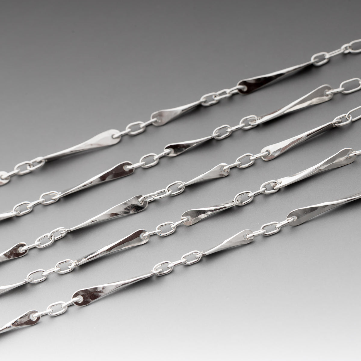 A close-up of handmade sterling silver chain bracelets featuring elongated, polished teardrop-shaped links connected by small oval links, displayed diagonally on a smooth gray gradient background.