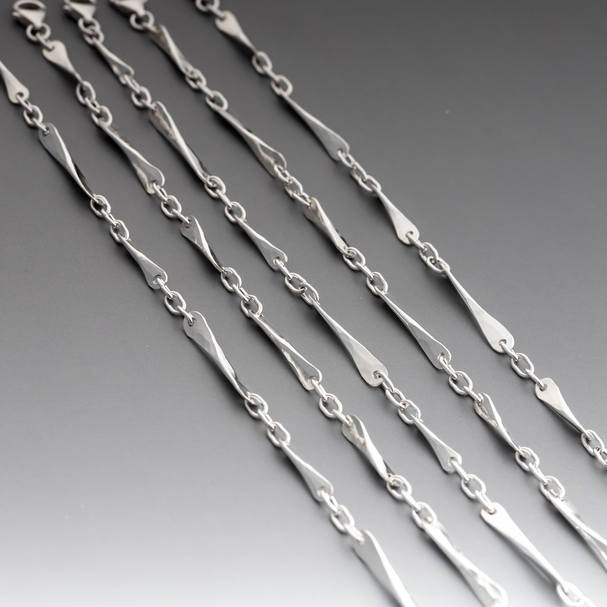 A close-up of handcrafted sterling silver bracelets featuring elongated, polished teardrop-shaped links connected by smaller oval links, set against a dark gray gradient background.