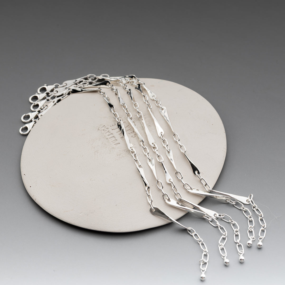 A set of polished hand forged sterling silver chain bracelets with lobster clasp ends, draped over a round beige display stand against a gray gradient background.  The links feature a hand hammered texture for a unique artisan style