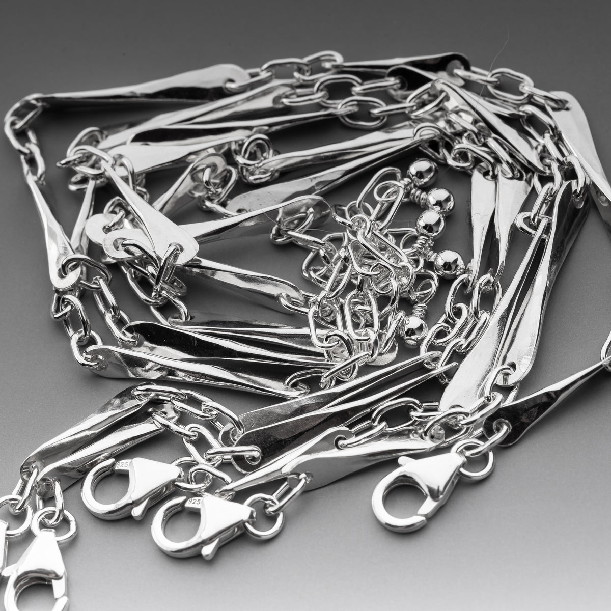 A close-up of handmade hammered bar bracelets with flat rectangular links, lobster clasp ends, and a small cluster of bead details, arranged on a neutral gray background.  These sterling silver bracelets are unique and one of a kind