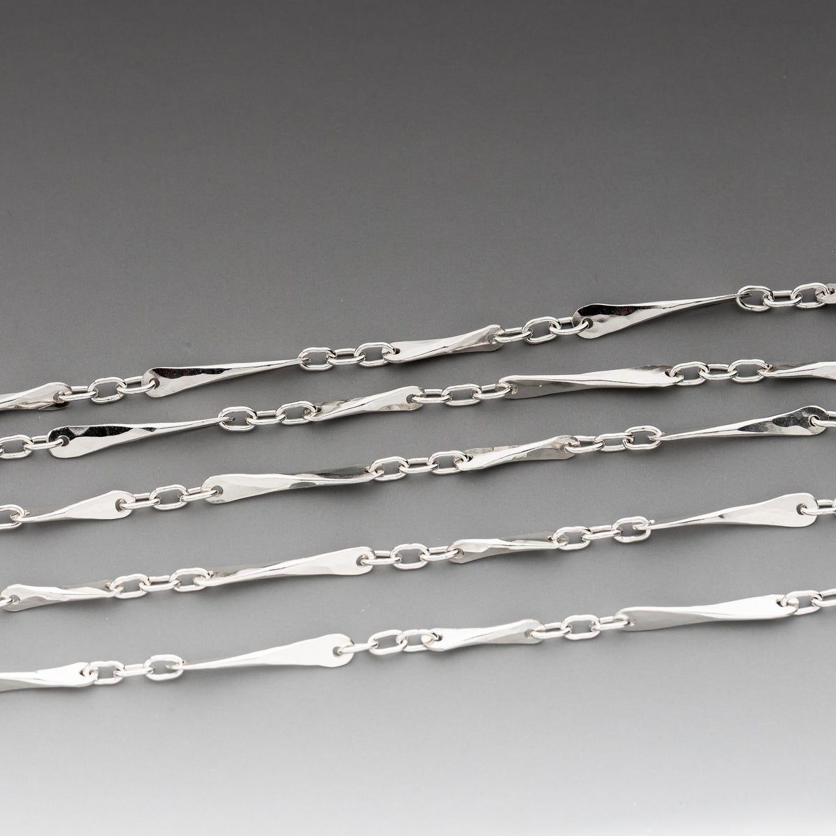 A close-up of handmade sterling silver chains with elongated, tapered oval links arranged diagonally on a smooth gray gradient background.