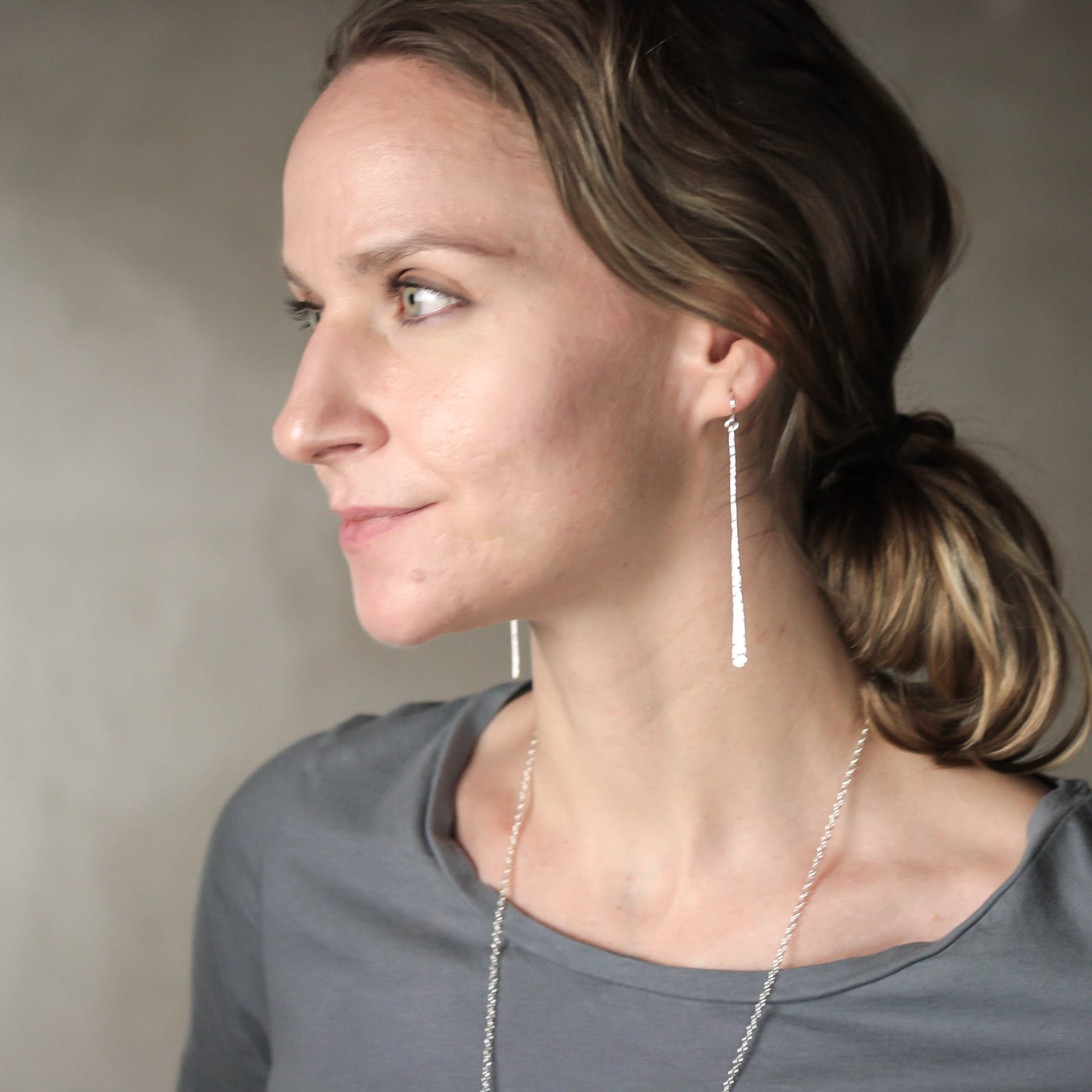 A woman in profile wearing a long, delicate silver drop earring and a matching silver chain necklace, set against a neutral gray background. Handmade by Lizardi Jewelry 