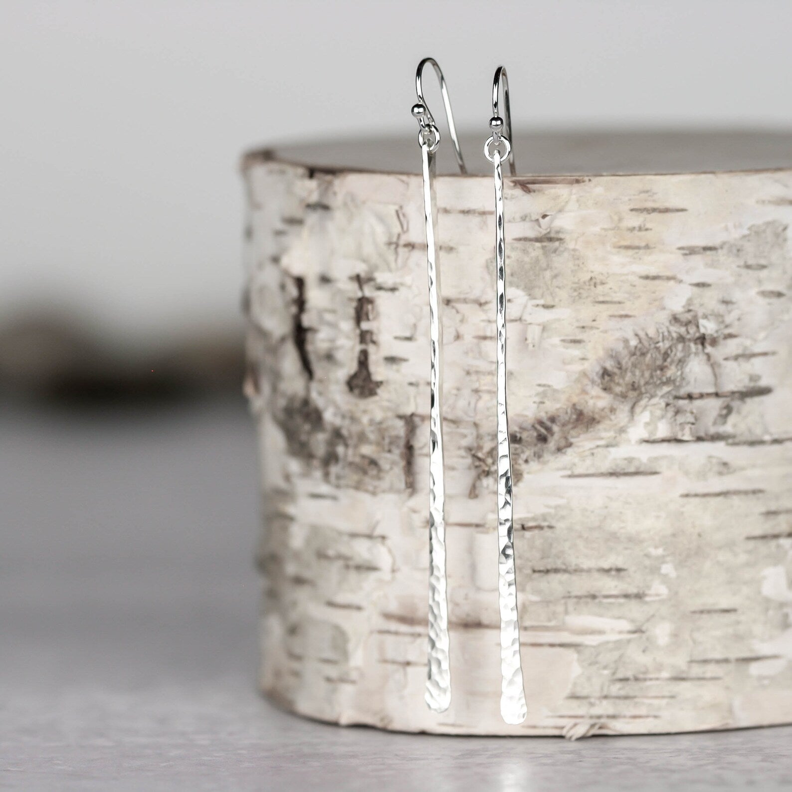 Two long, silver earrings hanging from a birch log on a neutral gray background. Handmade by Lizardi Jewelry 