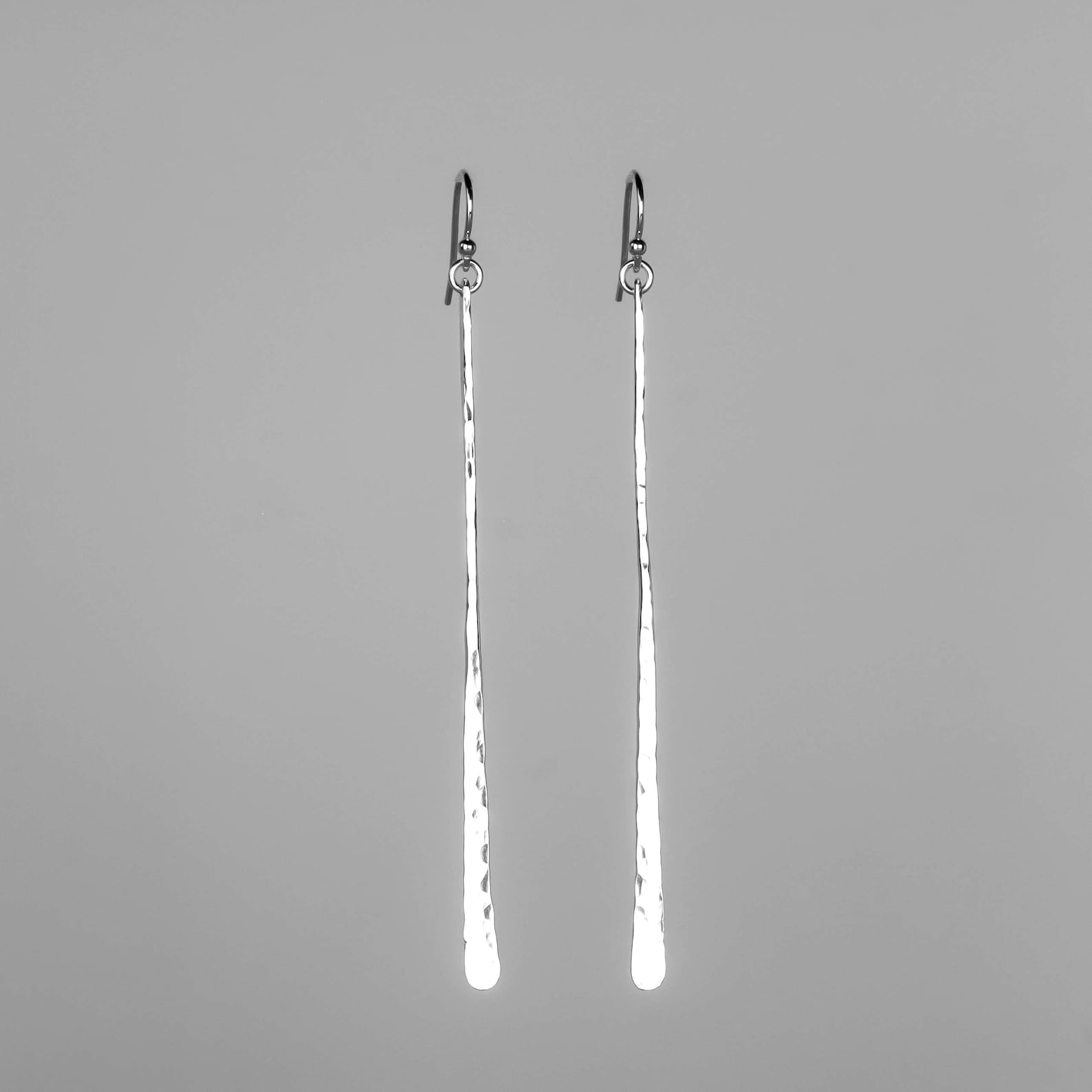 A pair of long, hammered silver bar earrings with fishhook backs, displayed against a solid gray background. Handmade by Lizardi Jewelry 