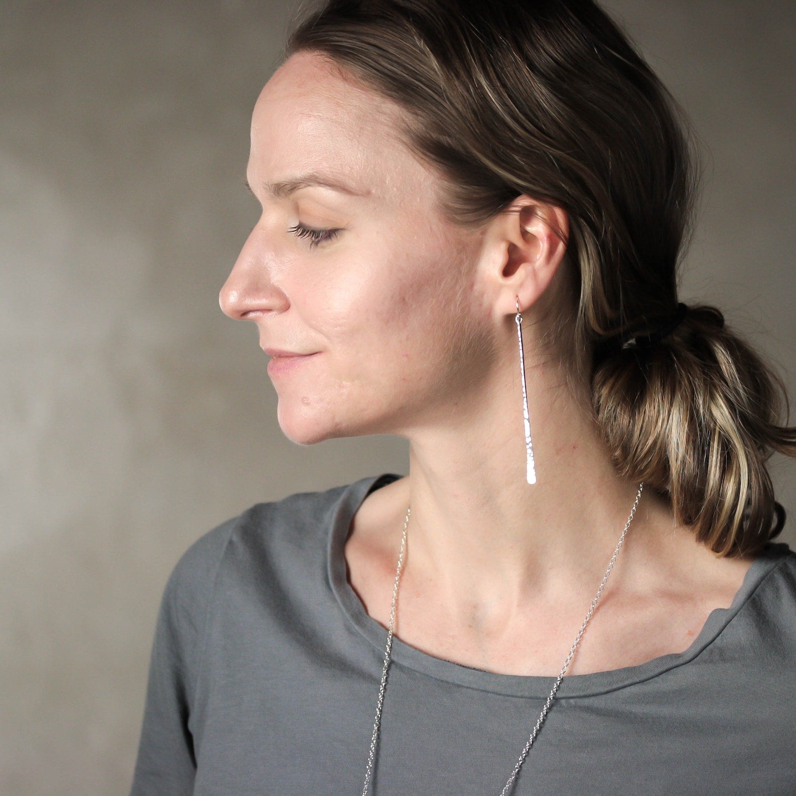 A woman in profile wearing a long, sterling silver earring and a  delicate silver necklace, set against a neutral gray background. Handmade by Lizardi Jewelry 