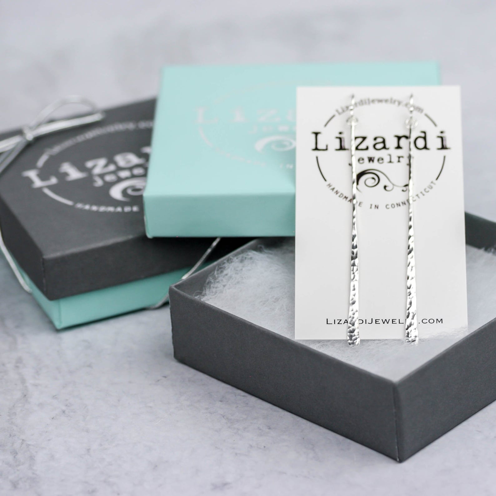Silver drop earrings displayed on a white Lizard Jewelry card, set in an open dark gray gift box with branded boxes behind it on a light stone surface.