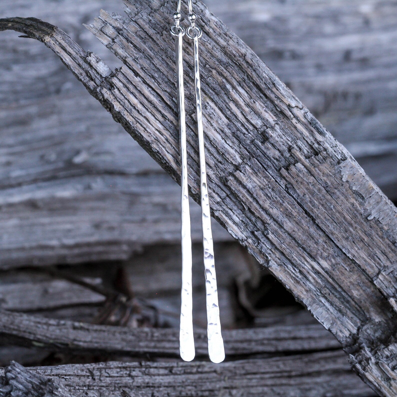 A pair of long, hammered silver bar earrings hanging from a weathered piece of wood against a blurred rustic wood background. Handmade by Lizardi Jewelry 