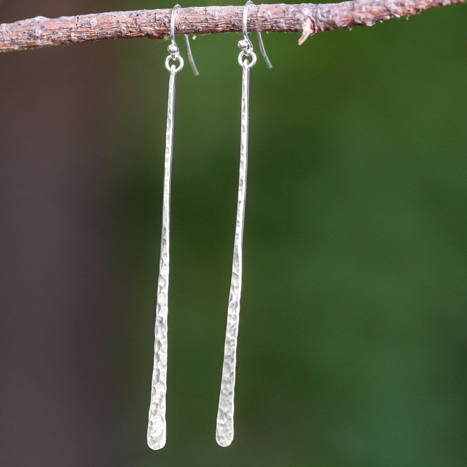 A pair of long, hammered silver drop earrings hanging from a small brown twig against a blurred green and brown outdoor background. Handmade by Lizardi Jewelry 