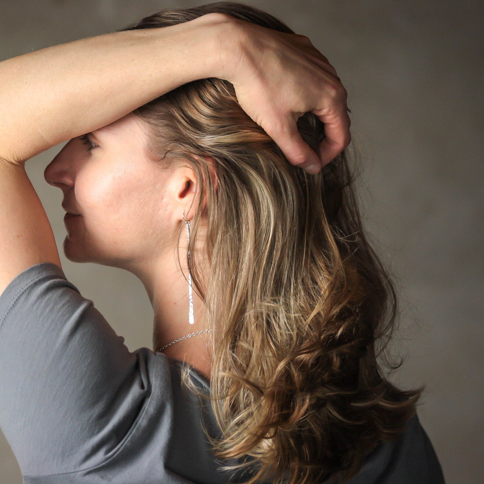 A woman with shoulder-length wavy brown hair wearing a delicate silver drop earring posing with her arm raised against a neutral gray background. Handmade by Lizardi Jewelry 