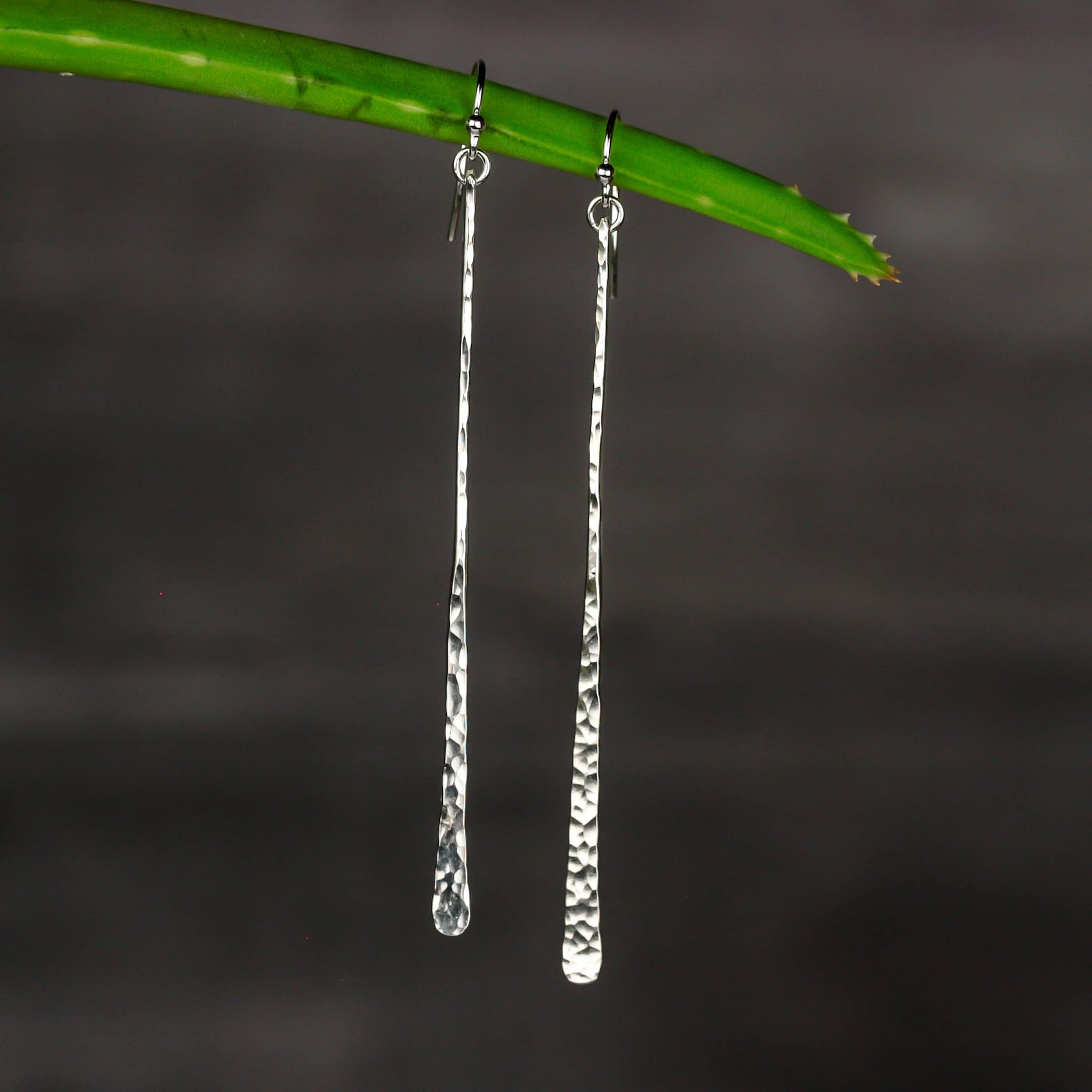 A pair of long, hammered silver earrings hanging from a green plant stem against a dark, blurred background. Handmade by Lizardi Jewelry 