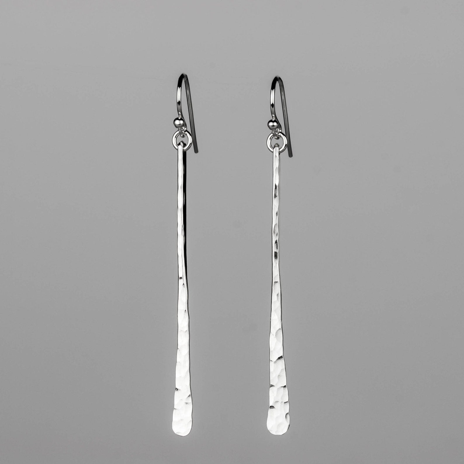 A pair of sterling silver drop earrings with a hammered finish and fishhook backings, displayed against a solid gray background. Handmade by Lizardi Jewelry