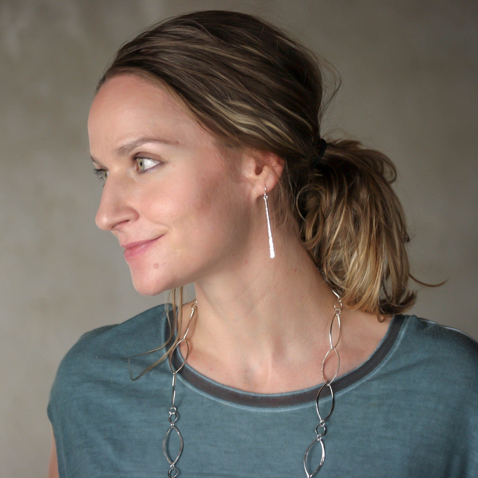 A woman with light brown hair in a low ponytail is shown in profile wearing a long, delicate sterling silver dangle earring and a matching long silver necklace with interlocking oval links. Handmade by Lizardi Jewelry 