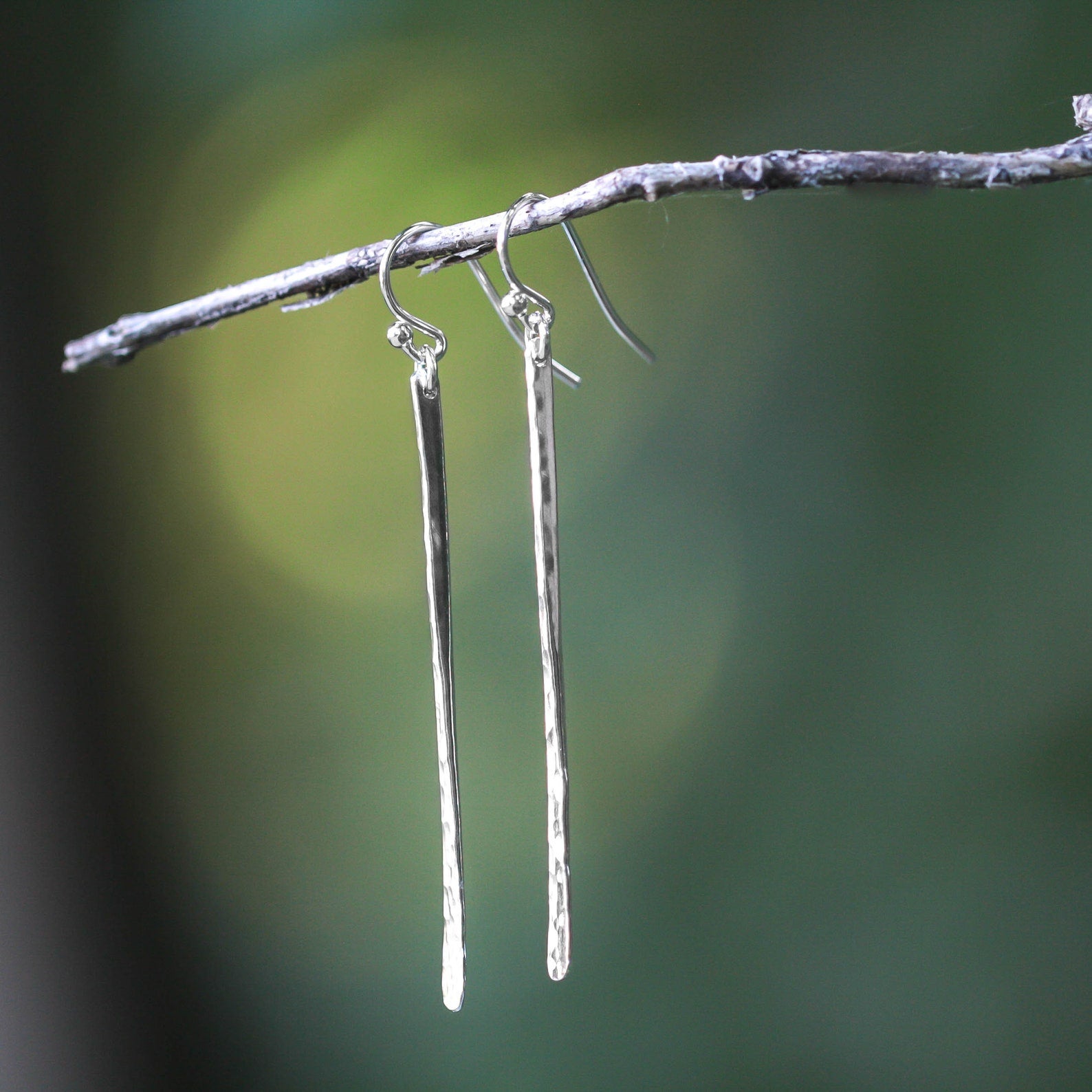 A pair of long, hammered silver drop earrings hanging from a thin branch against a soft, blurred green and dark background. Handmade by Lizardi Jewelry 