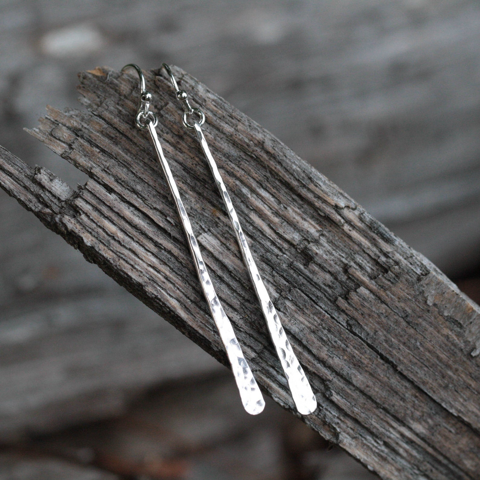 A pair of long, skinny silver drop earrings with fishhook backs, resting on a piece of weathered gray wood. Handmade by Lizardi Jewelry 