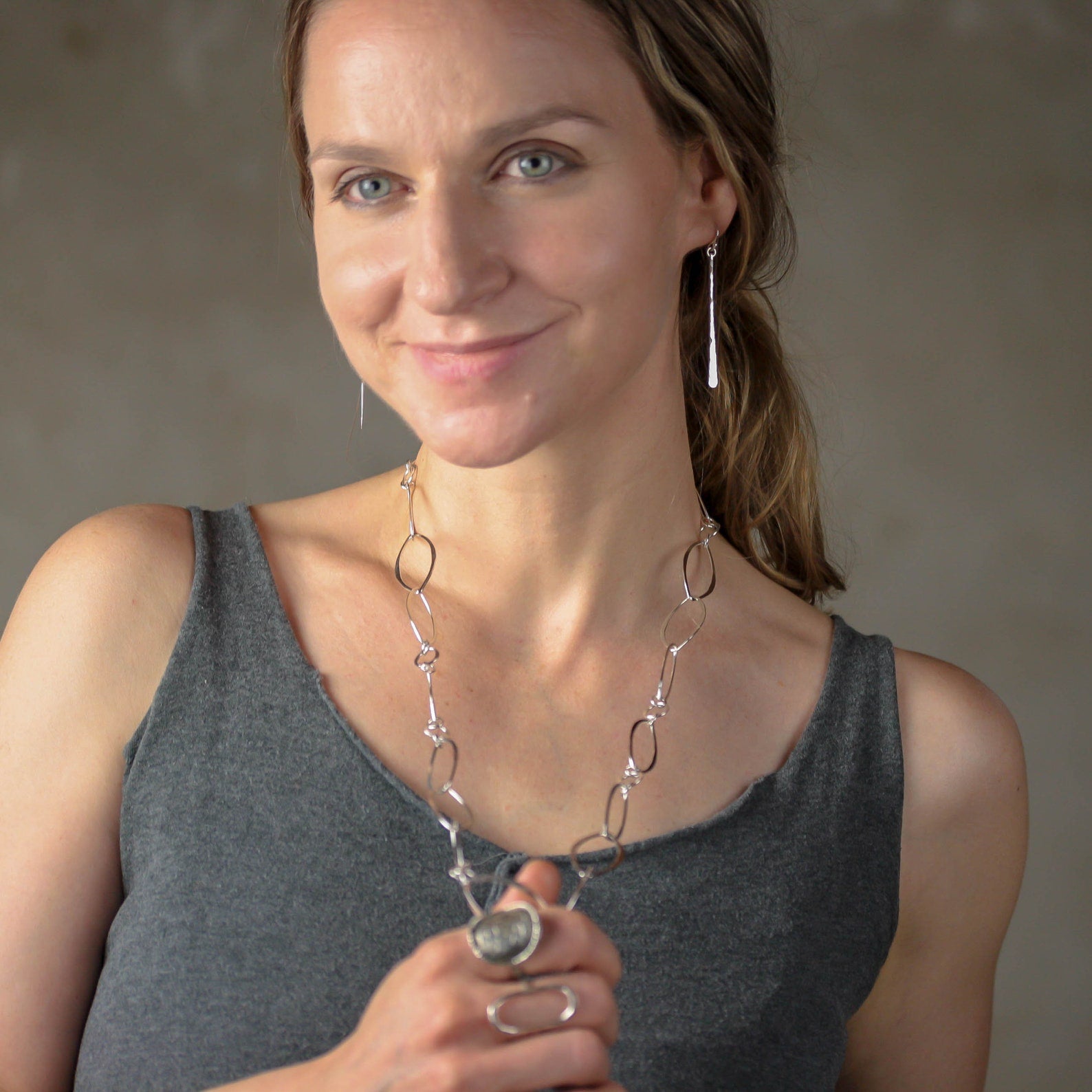 A woman wearing a silver link necklace, a matching silver drop earring, and silver rings, wearing a gray tank top against a neutral gray background. Handmade by Lizardi Jewelry 