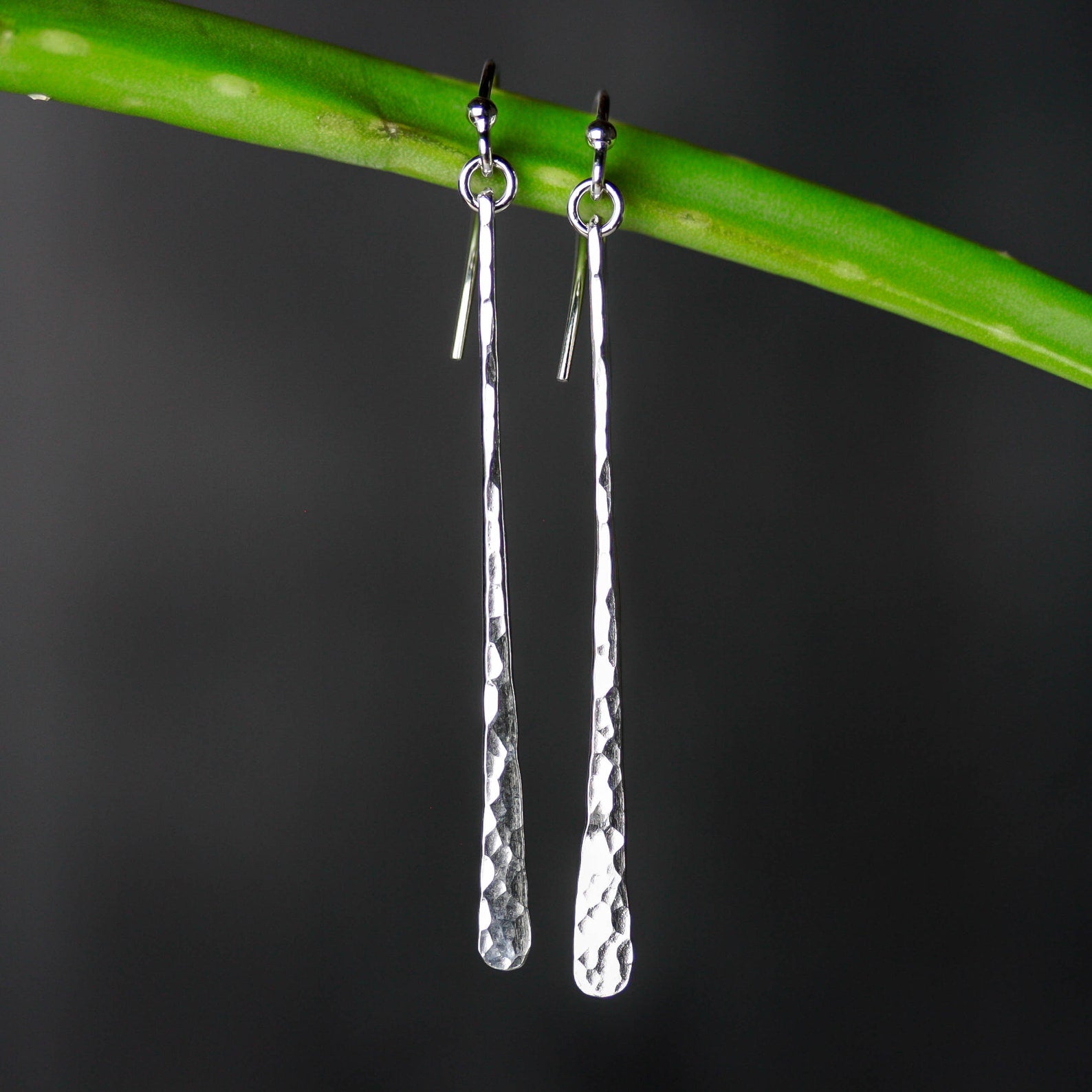 A pair of sterling silver earrings with a hammered texture hanging from a green plant stem against a dark gray background. Handmade by Lizardi Jewelry 