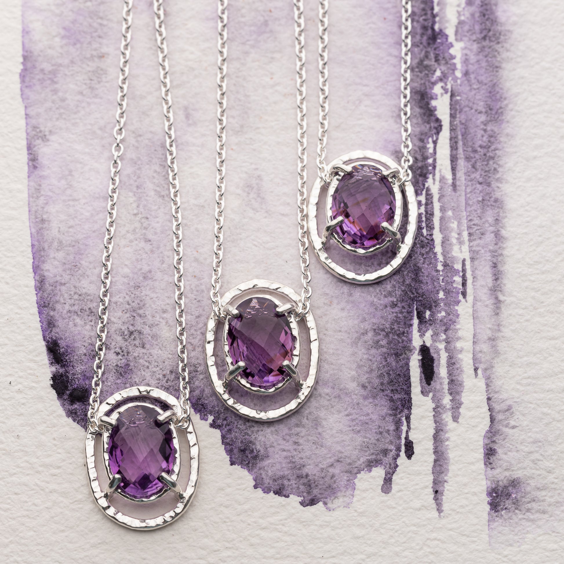 Three silver necklaces with purple amethyst gemstones on a watercolor purple and gray background