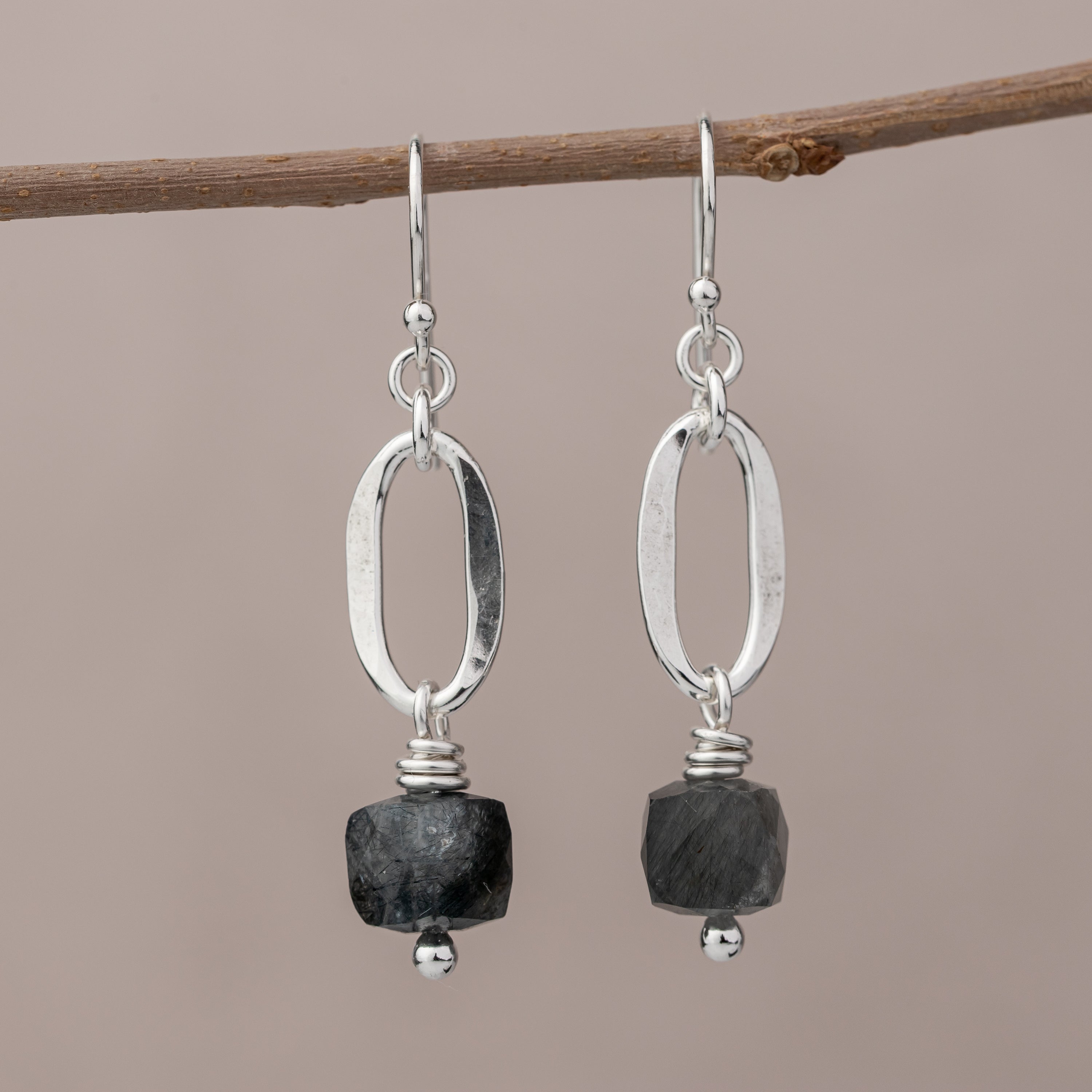 Handmade sterling silver earrings with dark quartz stones hanging on a branch against a beige background