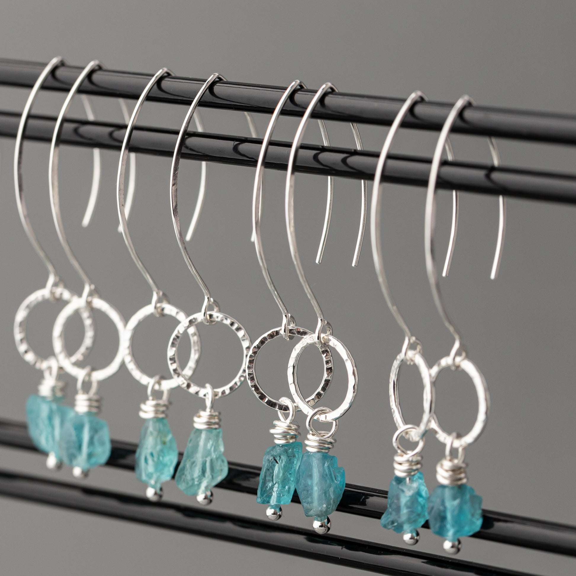 Silver hoop earrings with blue apatite raw stones on a black rod against a gray background