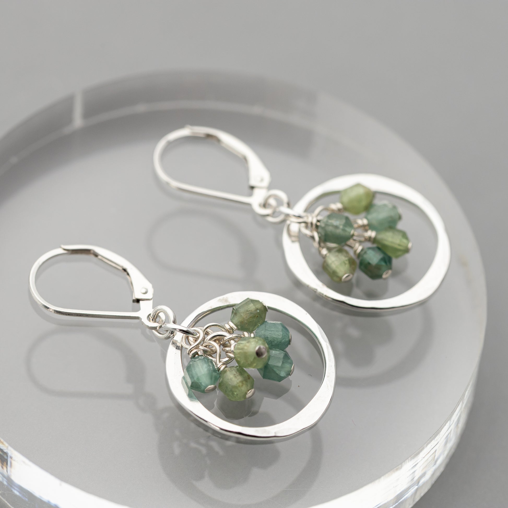 Sterling silver hammered circle earrings with green gemstones clustered together in the center on a reflective surface