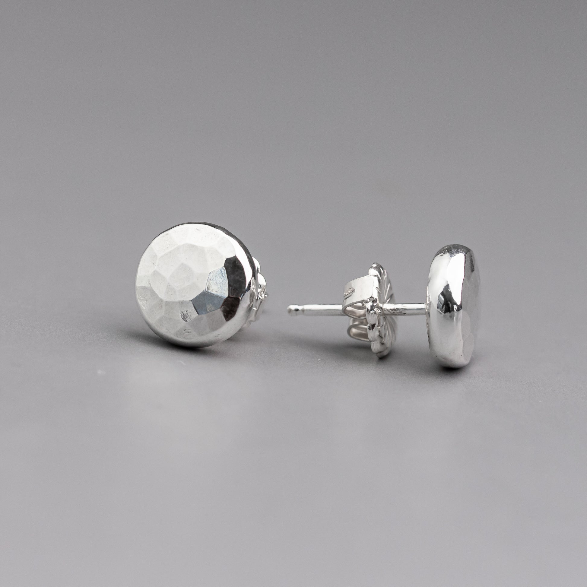 a pair of sterling silver studs shown with one on profile to show the post and ear nut and the other straight on to highlight the unique hammer texture on the surface of the earrings