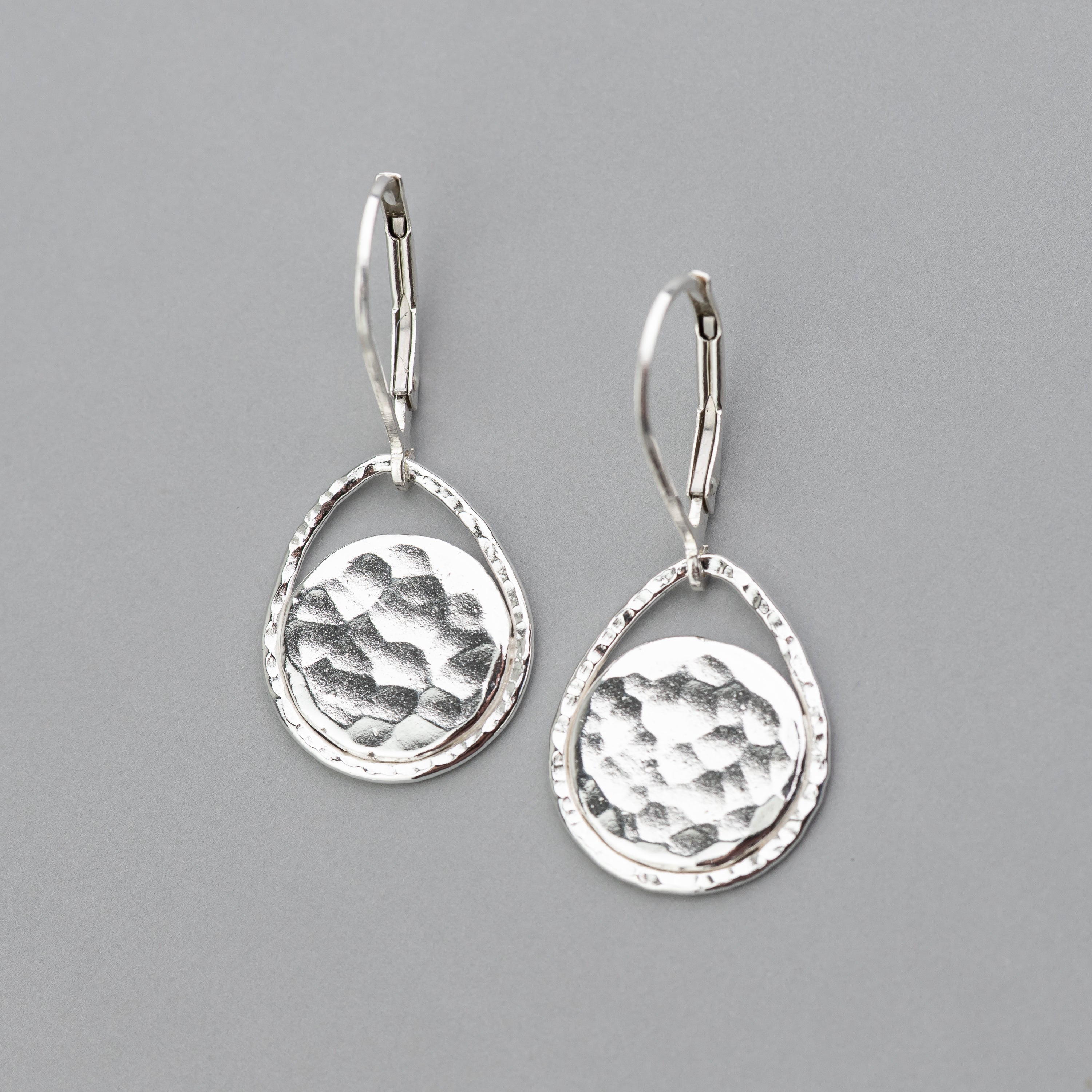 Handmade sterling silver lever back earrings shown against a gray background. The dangle earrings feature hand hammer textured discs that are surrounded by an open teardrop shape to frame them beautifully. 